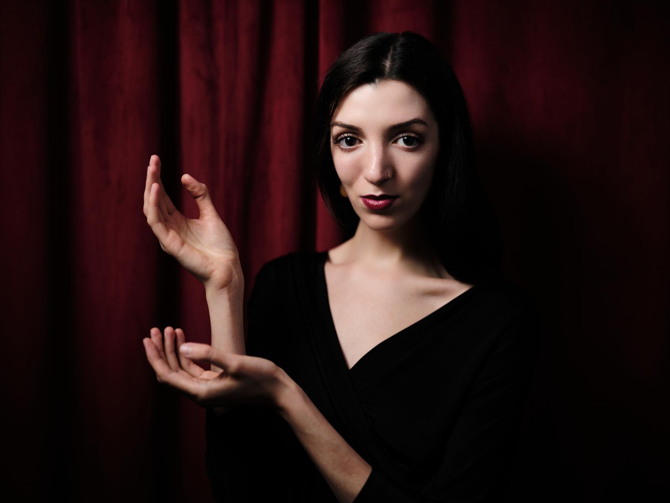 Studio portrait of a woman with dark hair in a black V-neck top, posing with raised hands against a deep red curtain backdrop under soft, directional lighting.