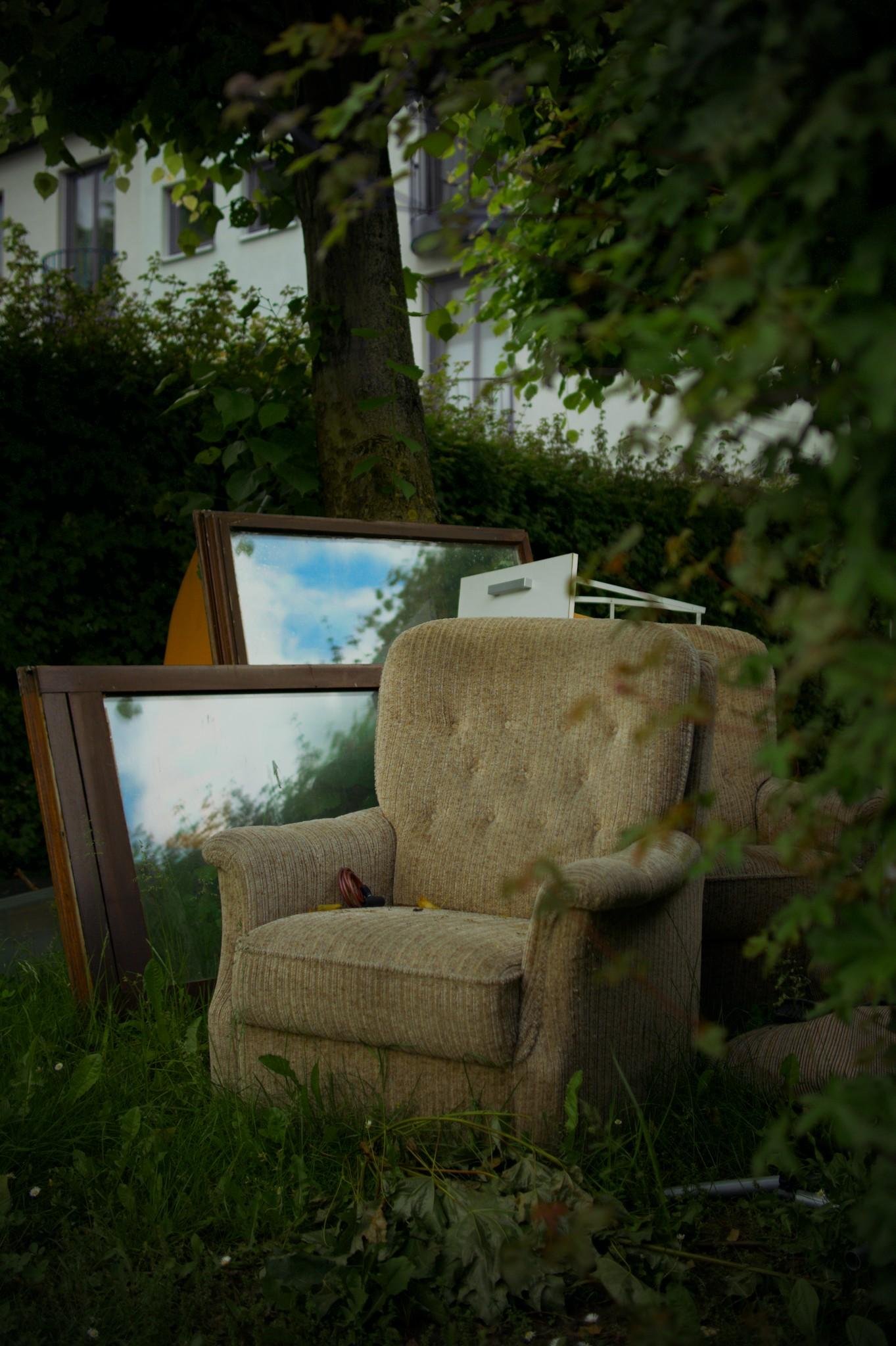 Beige upholstered armchair on grass under leafy trees, with several large framed mirrors and a small white shelf stacked behind it; apartment building windows visible in the background.