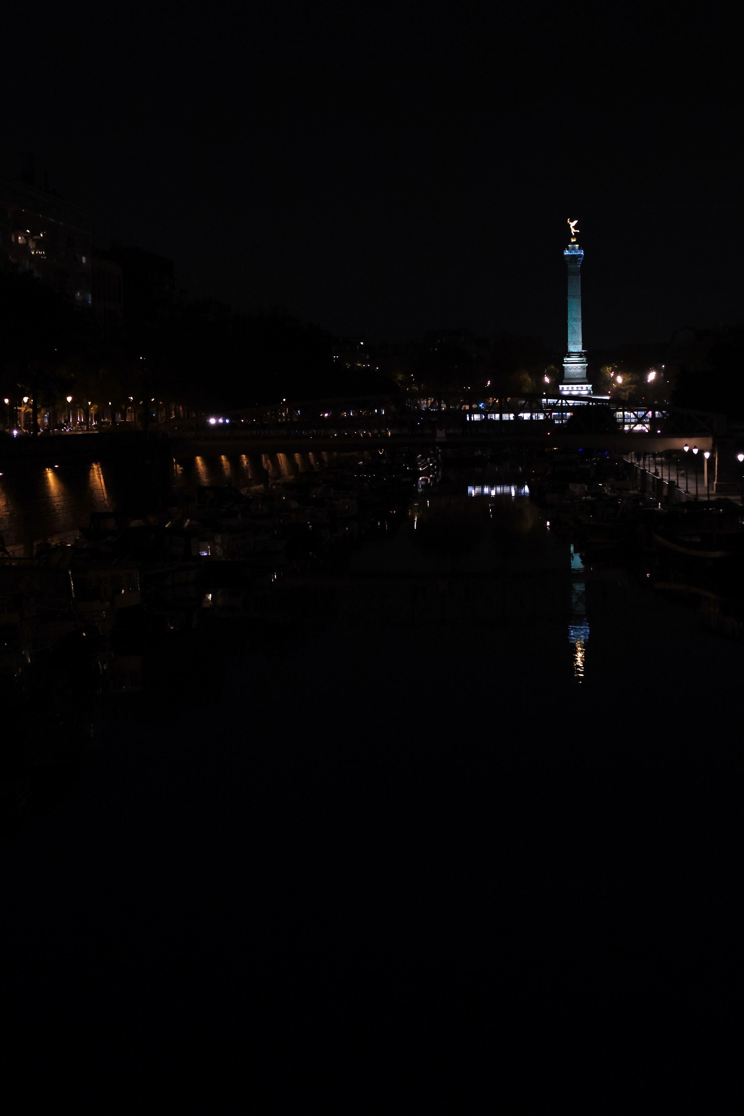Photo format portrait, de nuit, montrant au fond, le pilier de la place Bastille à Paris, se reflétant dans la Seine qui occupe la moitié basse de l’image. À gauche, des lampes oranges éclairent un quai de Seine vers le point de fuite de la photo