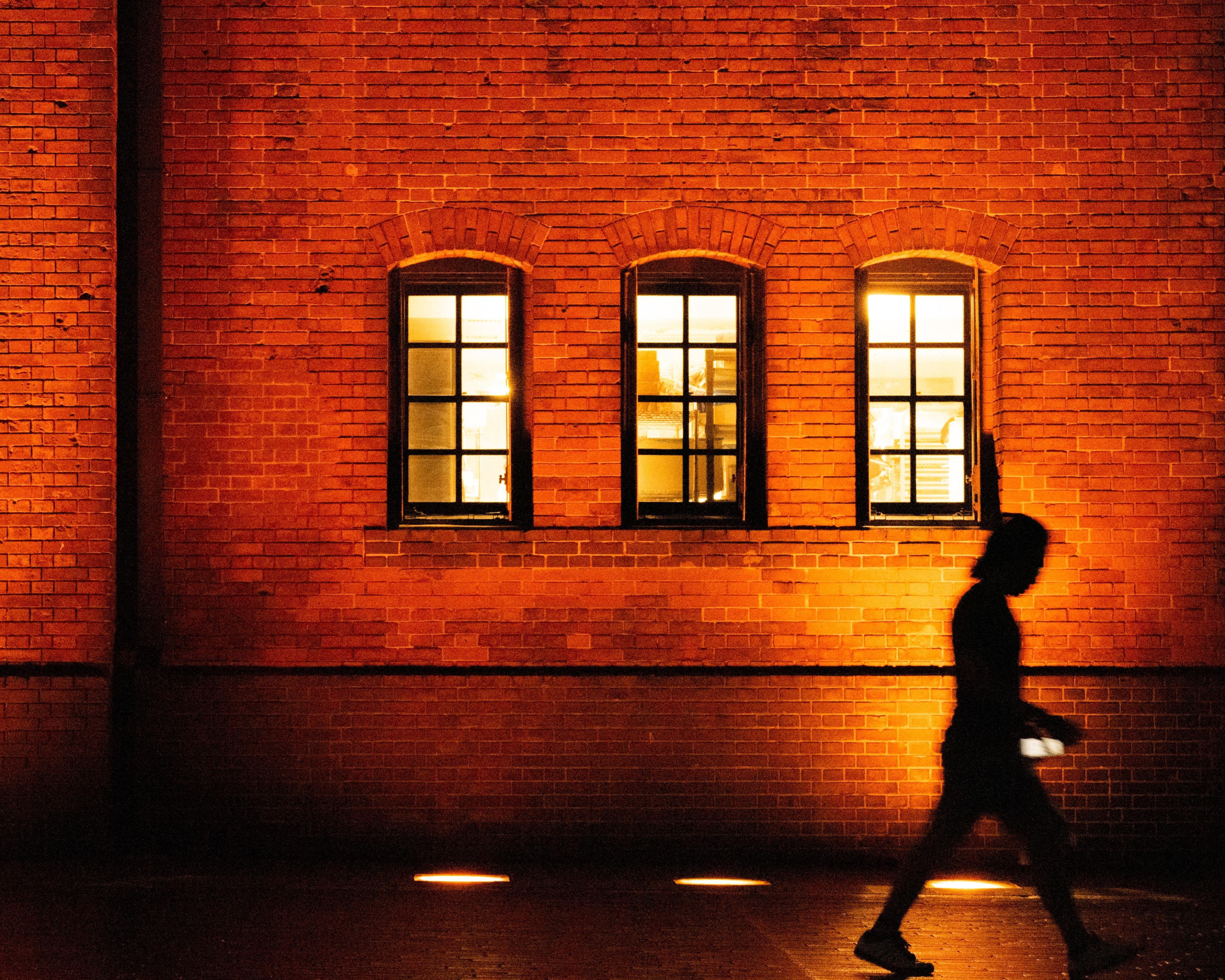 Brick wall with three arched windows, warmly lit from inside. Silhouette of a person walking in front, holding a phone, creating a contrast against the illuminated background.