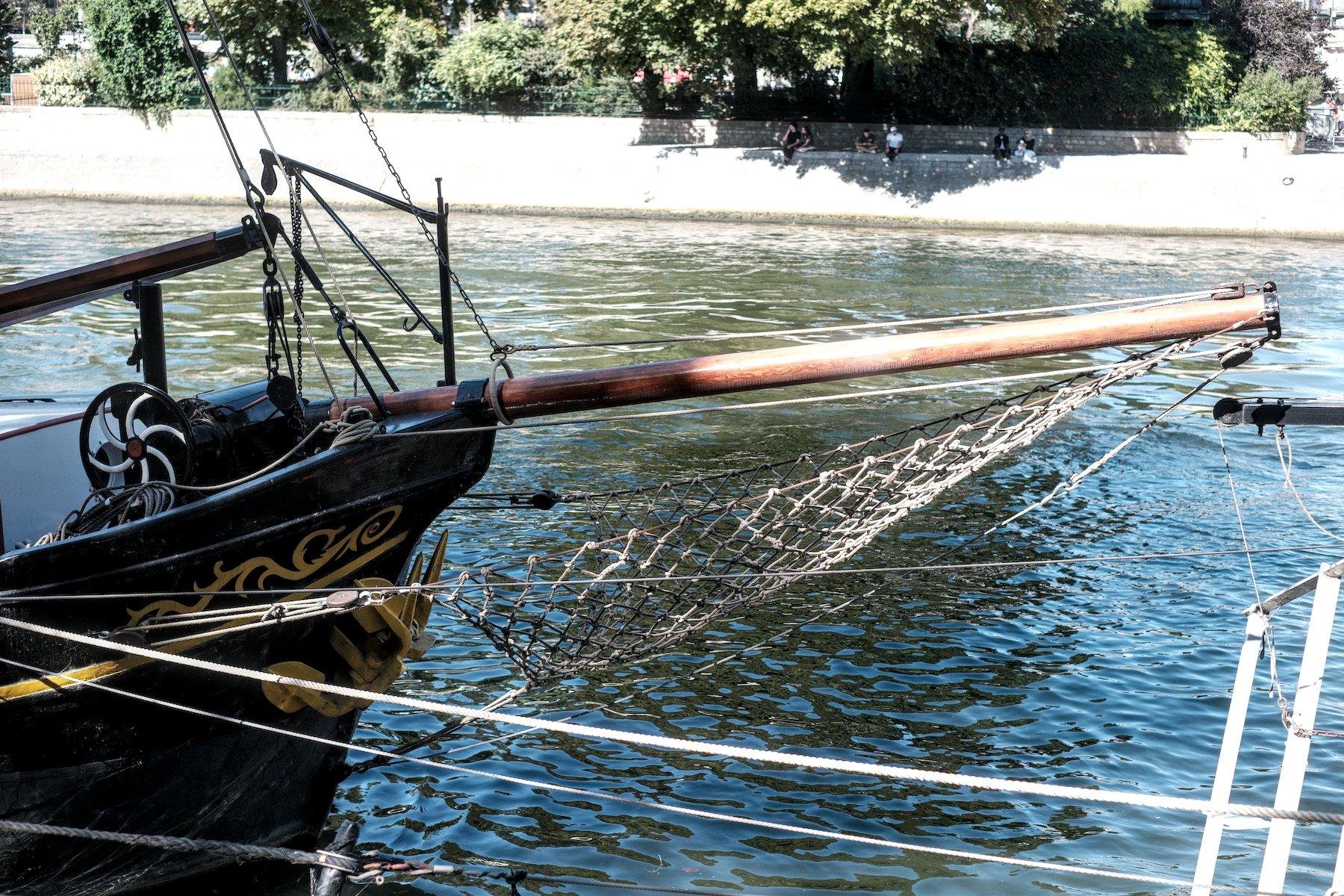 Photo horizontale. Proue noire décorée de motif doré abstrait d’un bateau avec la figure de proue étant un poteau en bois depuis le bout duquel est accroché un filet qui est attaché au bateau plus bas, formant un motif de voile pendante