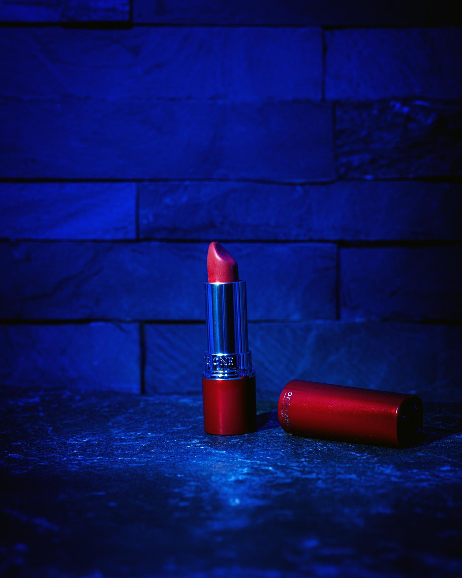 The image shows a tube of red lipstick with its cap placed beside it. The background and surroundings have a bluish tone.