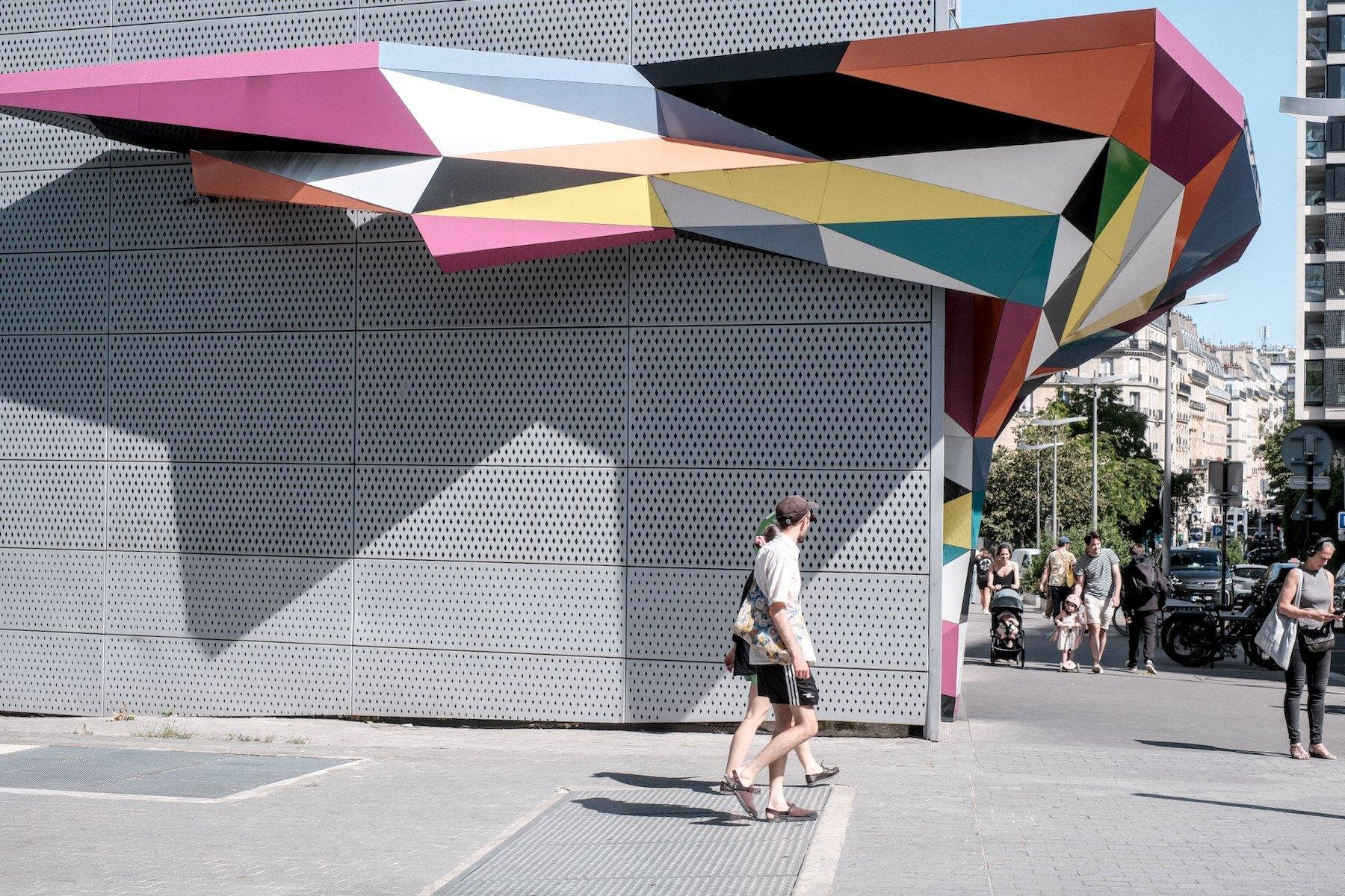 Photo horizontale. Sur un bâtiment gris, une espèce de toit de préaut de forme irrégulière, tout en angles, accentués par les motifs triangulaires de couleurs diverse et jamais consécutives, enveloppant l’angle du bâtiment. Des gens passent devant sous un soleil éclatant