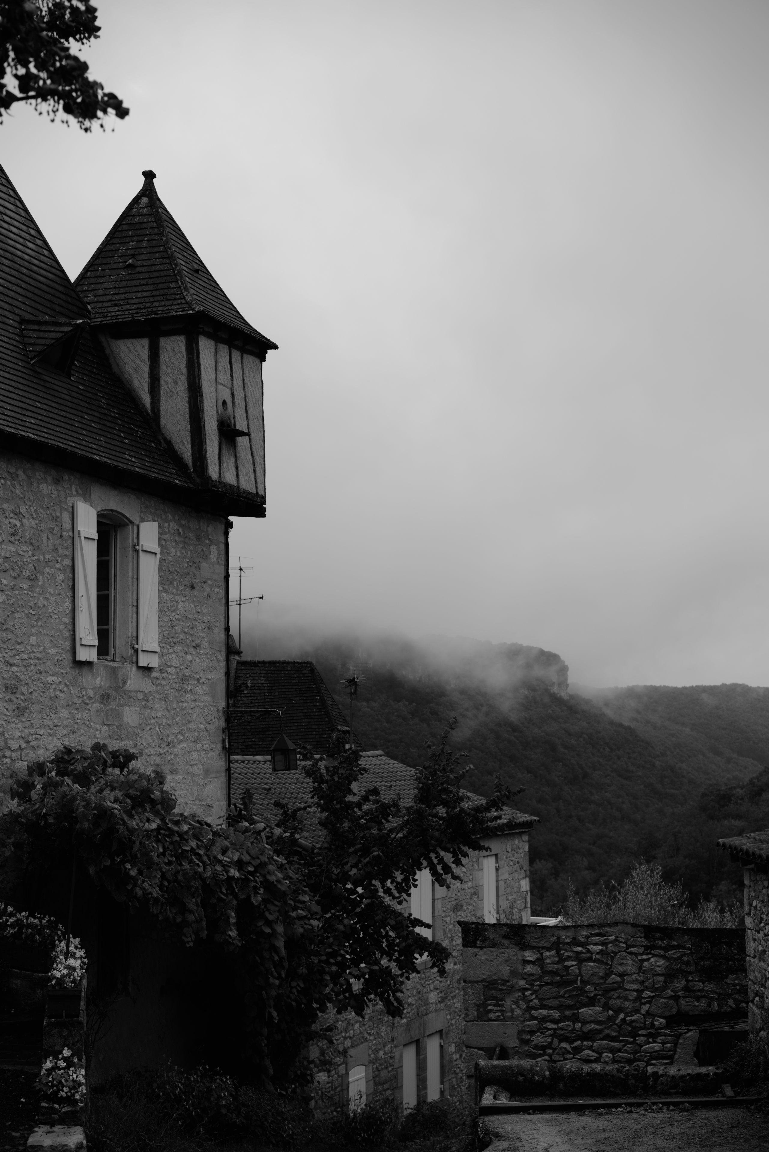 Photo format portrait en noir et blanc montrant à gauche un bout de maison en pierre avec une mini tour au toit pointu, avec un paysage de montagne embrumée au fond