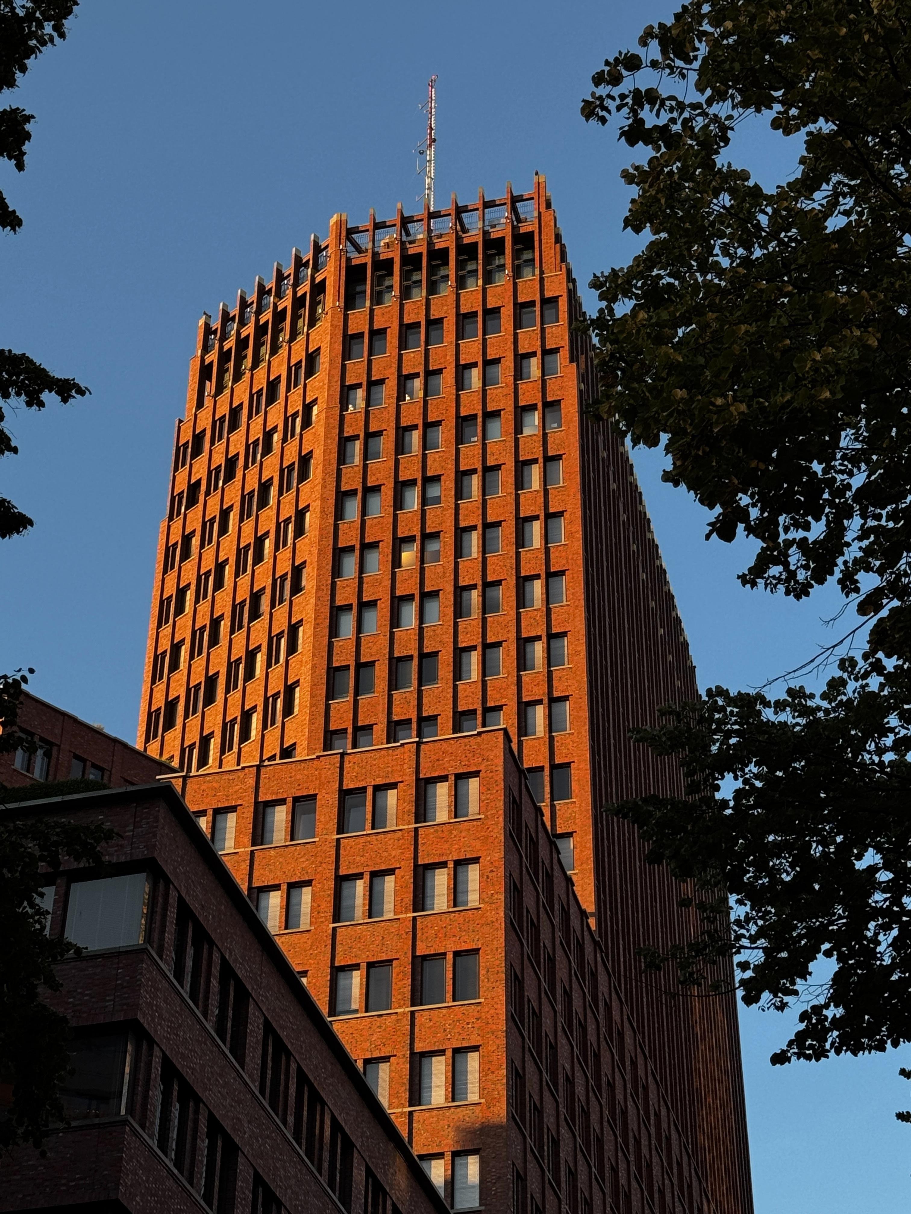 Tall, rounded brick building with vertical rows of windows and a spire, surrounded by smaller buildings and trees under a blue sky.