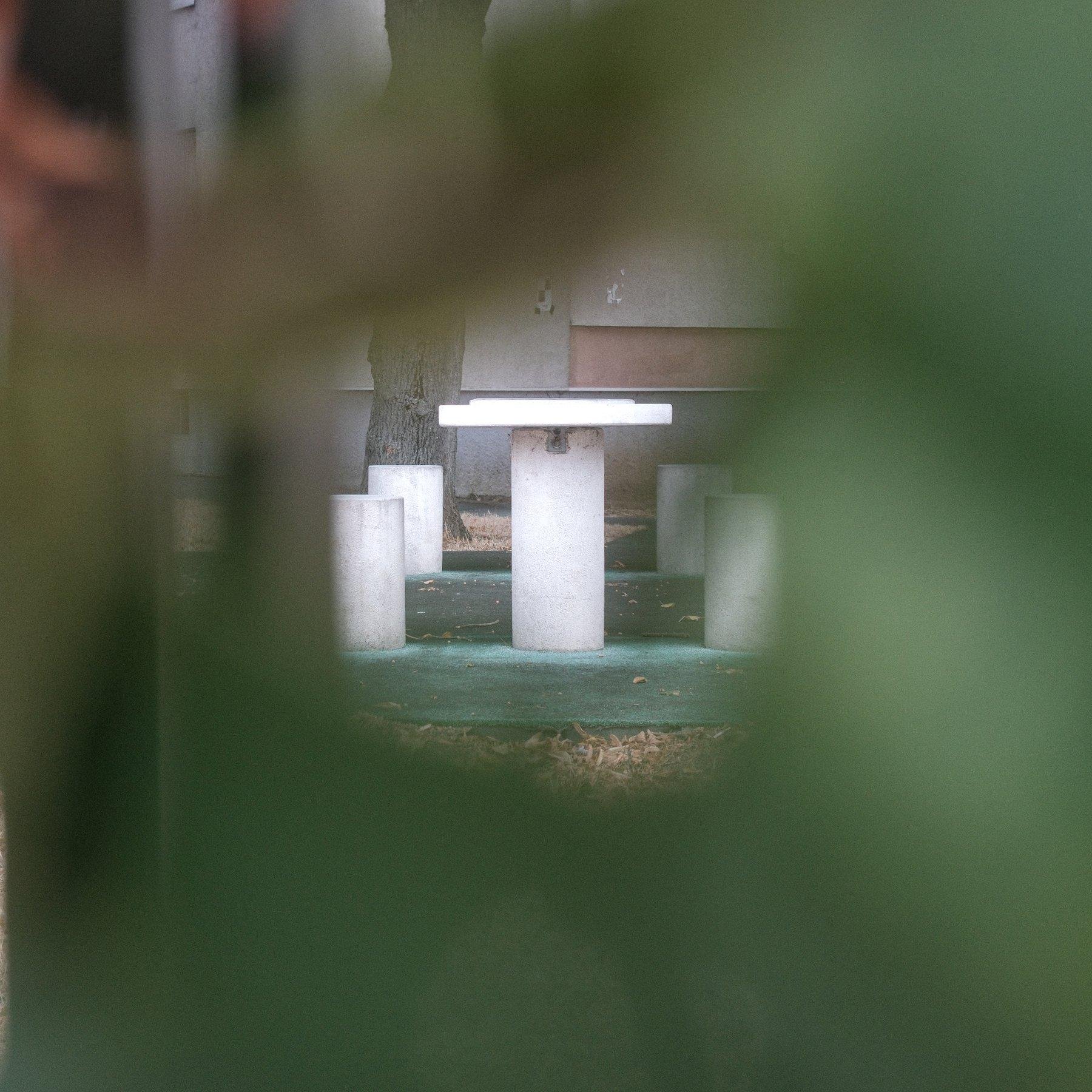 Photo carrée. Au premier plan, flou, de la végétation et au centre, entre les feuille, on voit une table blanche, de profile, sur un support blanc en béton et de chaque côté, un tabouret en béton blanc aussi. Personne n’est installé. On devine une seconde table derrière, comme en témoignent le set de deux autres tabourets en béton derrière