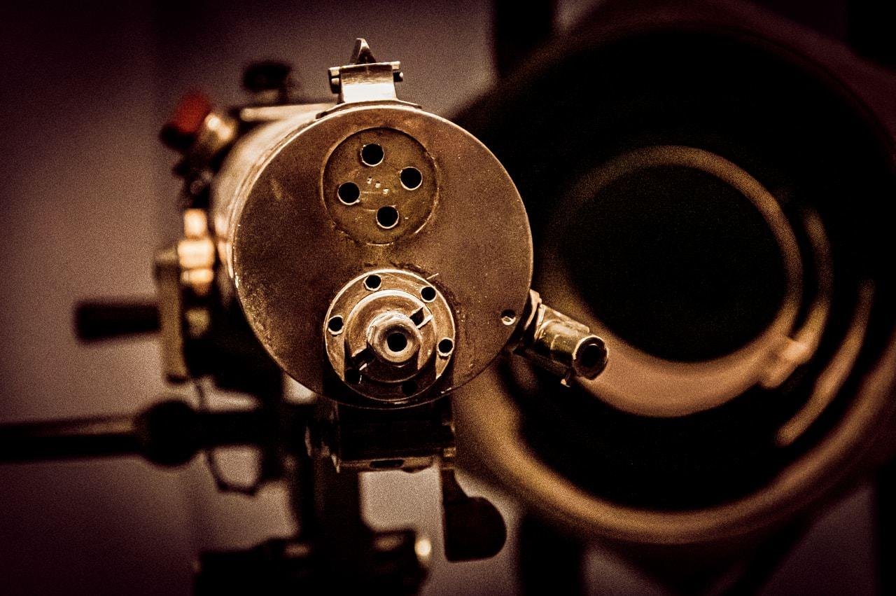 Antique machine gun with a cylindrical metal body, featuring multiple circular openings on the front. The image focuses on the projector head with blurred background elements suggesting an optical component, possibly a lens or reel. Dim lighting and sepia tones accentuate the vintage appearance.