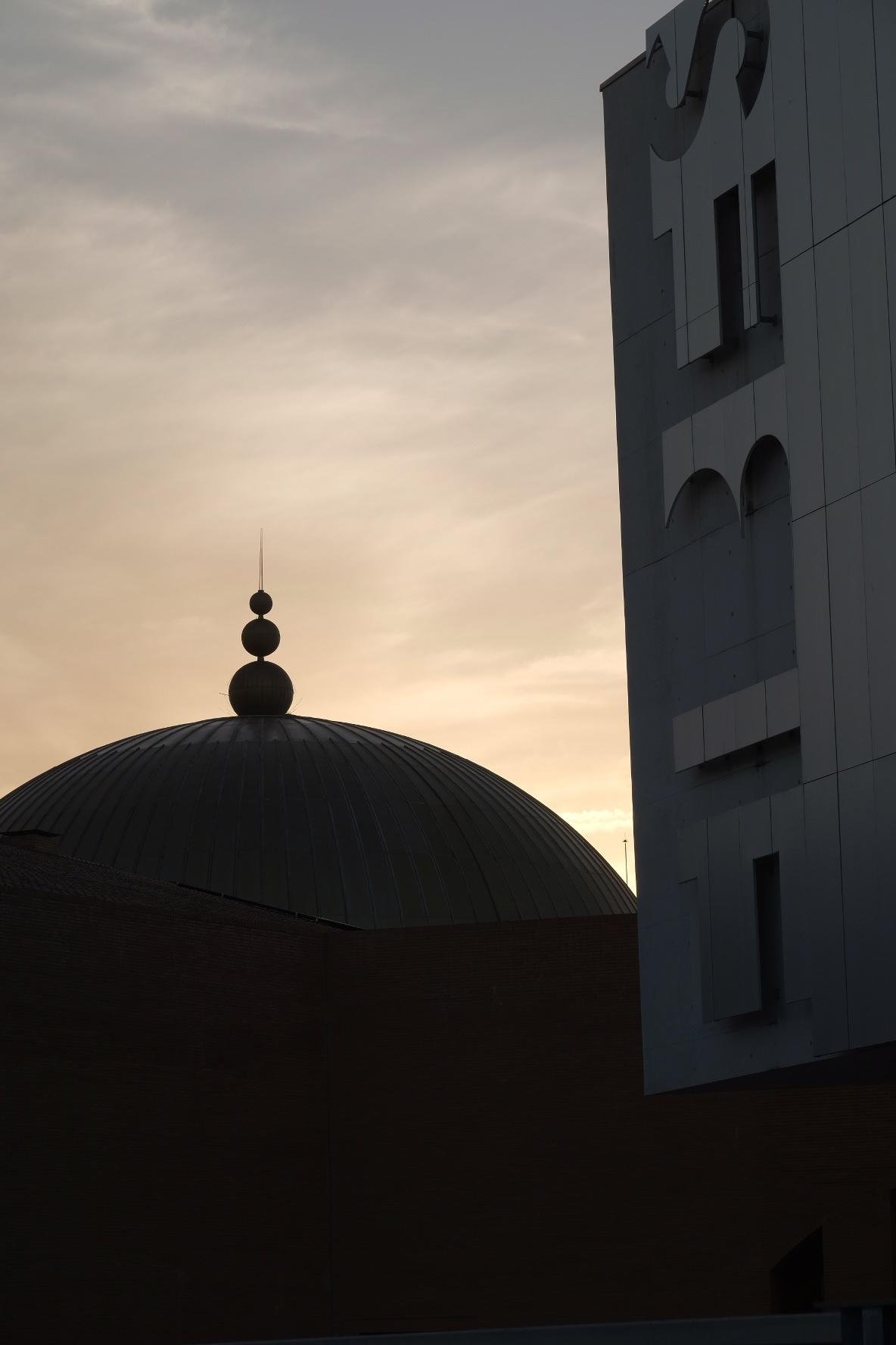 Dark dome roof topped by three stacked spheres silhouetted against a cloudy sunset sky, beside a tall modern building with large cutout shapes on its facade, spelling FIBES.