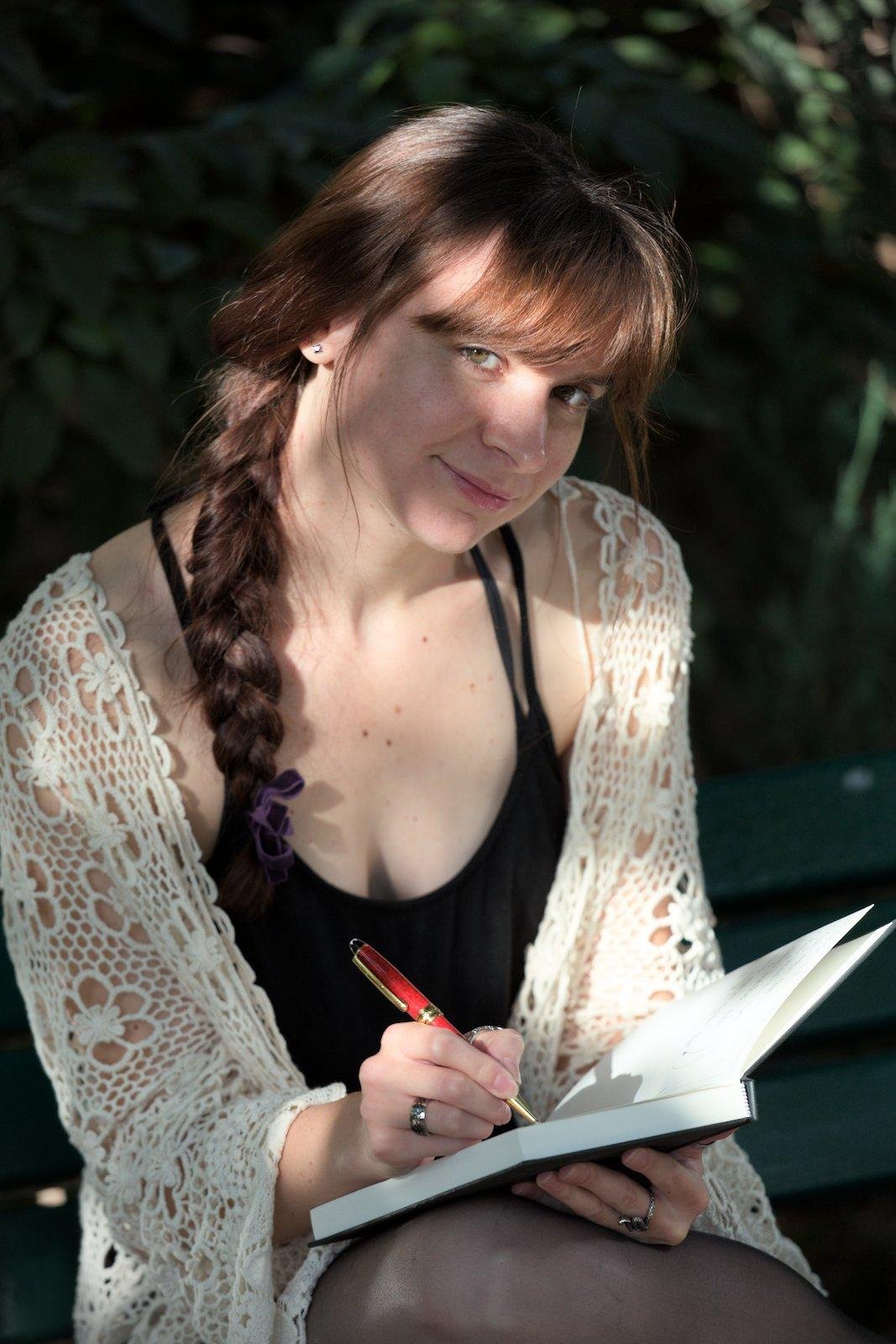 Photo format portrait. Une jeune femme châtain au cheveux longs coiffés en une longue natte qui pends devant son épaule droite. Elle porte un débardeur noir, par dessus duquel se trouve une espèce de cardigan en crochet blancs avec des motifs de fleurs, des collants noir. Elle tient un carnet de notes et un stylo rouge avec lequel elle était en train d’écrire avant de relever la tête vers la caméra avec un sourire et un regard doux. Le soleil perce des feuillages et l’éclaire de façon aléatoire