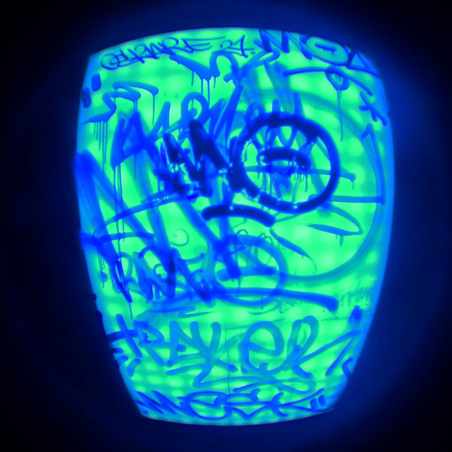 This image shows a neon blue-tinted empty illuminated advertising billboard covered in graffiti. The bright colours and abstract forms suggest a creative, urban art style. The image is taken through a fisheye lens.