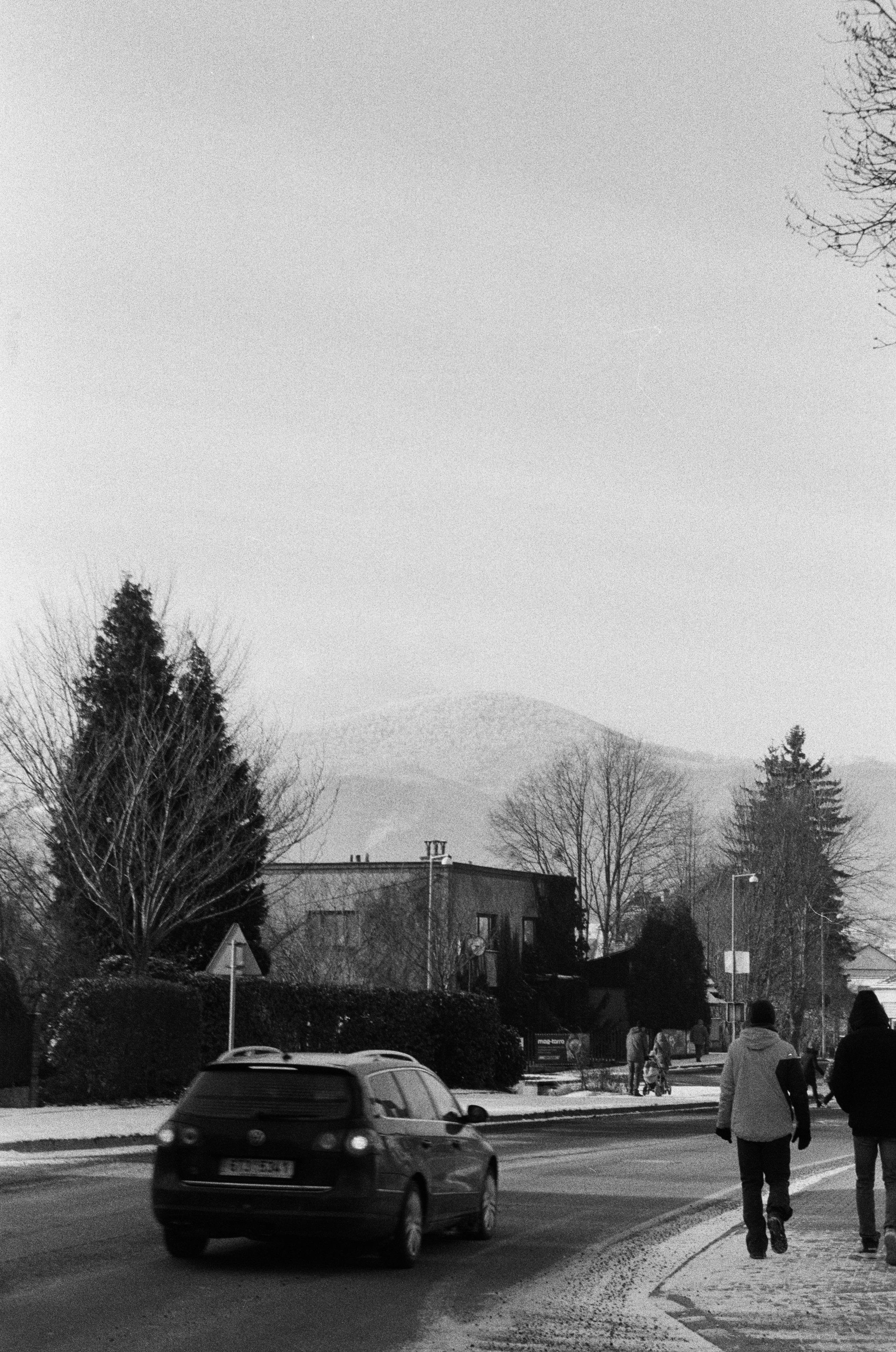 This black and white image shows a street scene. There is a car driving on the road, and people walking along the sidewalk. The background includes bare trees and a building. In the distance, there are hills or mountains visible. The scene appears to be in a colder season, possibly winter, as there seems to be snow or frost on the ground.