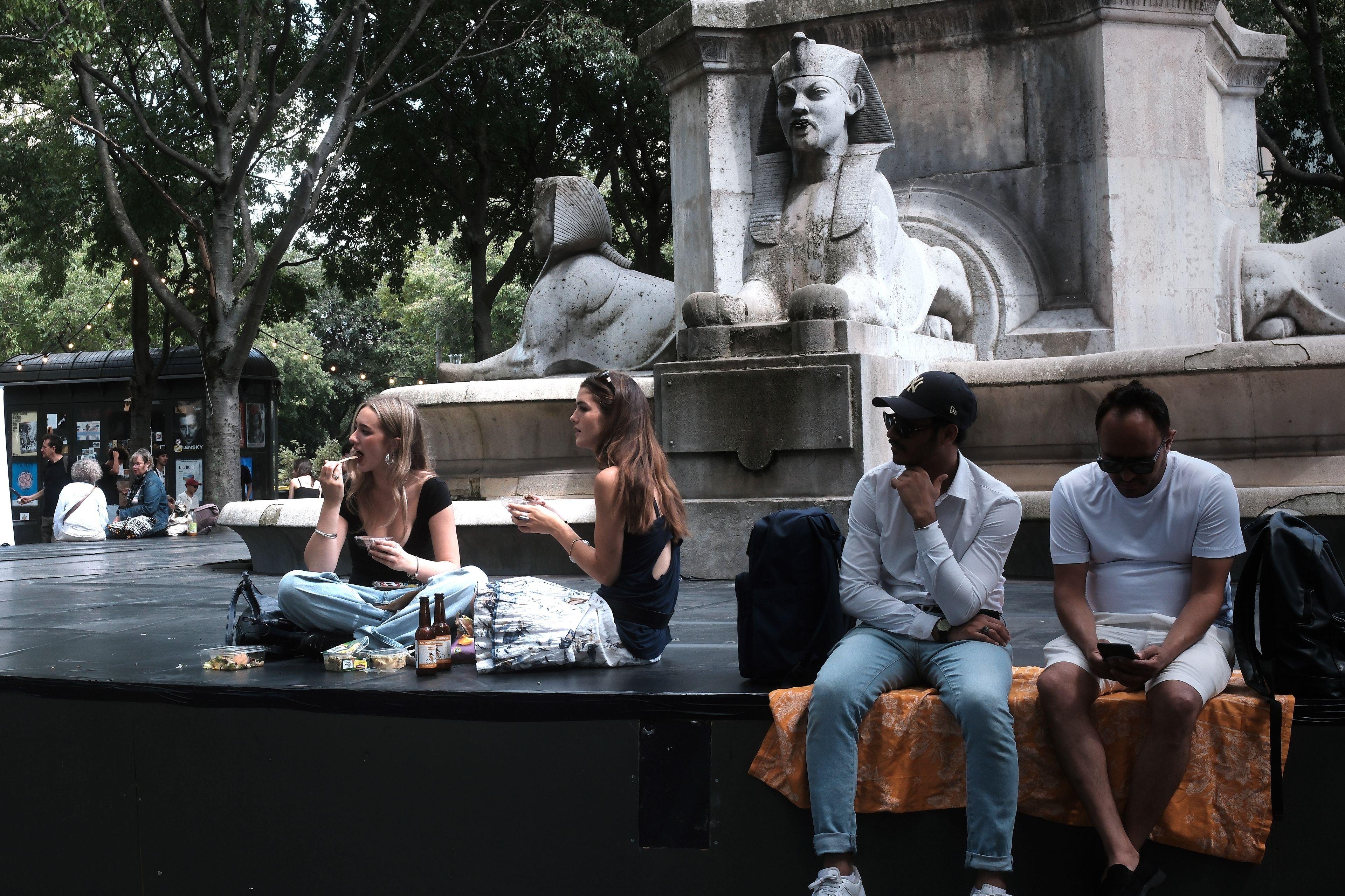 sur la gauche de la photo, 2 filles assises en tailleur devant une fontaine ornementée de sculptures de sphinx, mangent une glace en regardant dans la même direction hors cadre, des emballages de repas et deux bouteilles de bière se trouvent devant elles. Sur la droite, 2 hommes assis sur une serviette orange celui de gauche regarde aussi dans la même direction, celui de droite regarde son tél