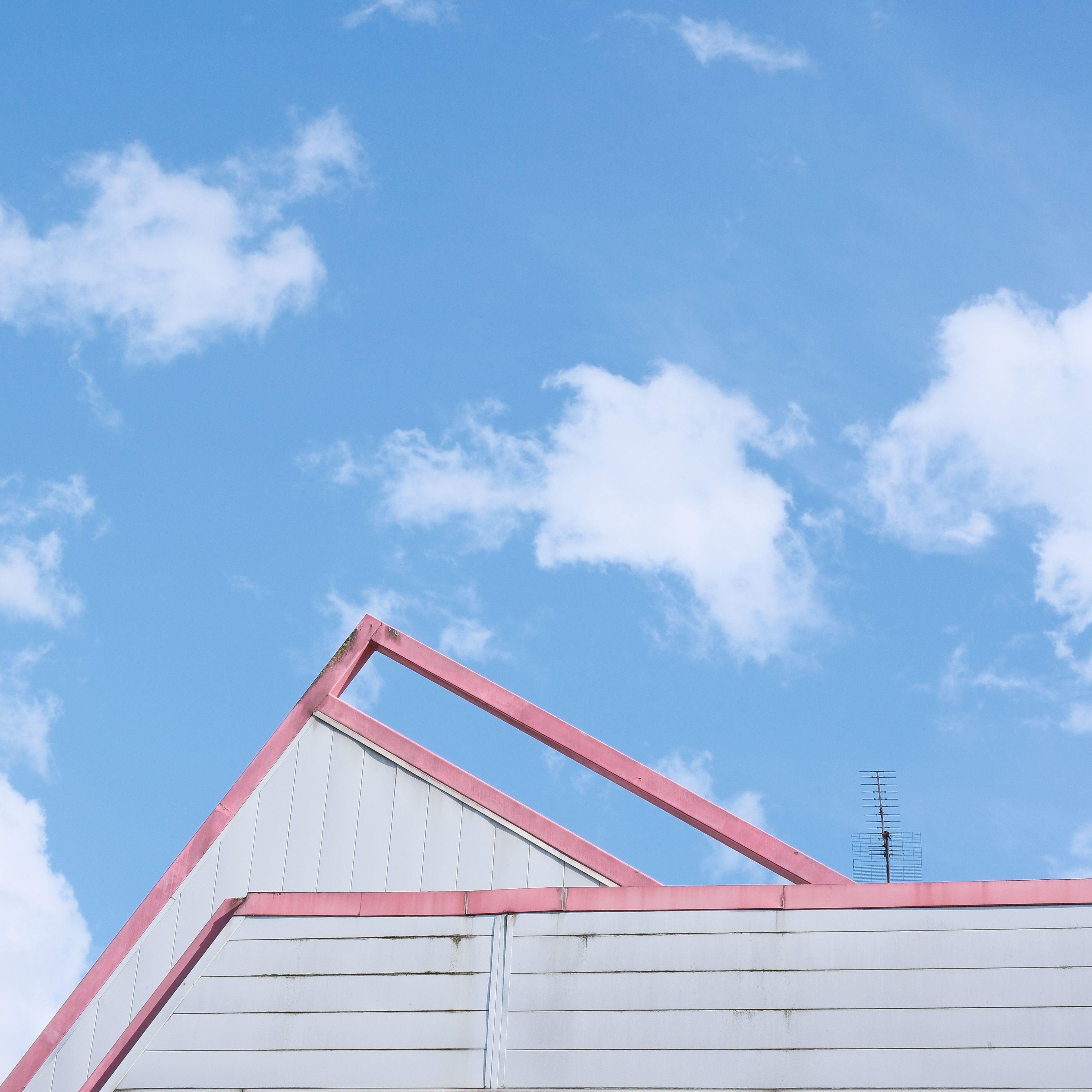 Photo carrée avec le ciel avec quelques nuage occupant les 3/4 de l’image. Le dernier quart en bas de la photo dépeint des formes géométrique par des murs et un toit gris dont les bords rouges soulignent leur délimitation avec le ciel bleu