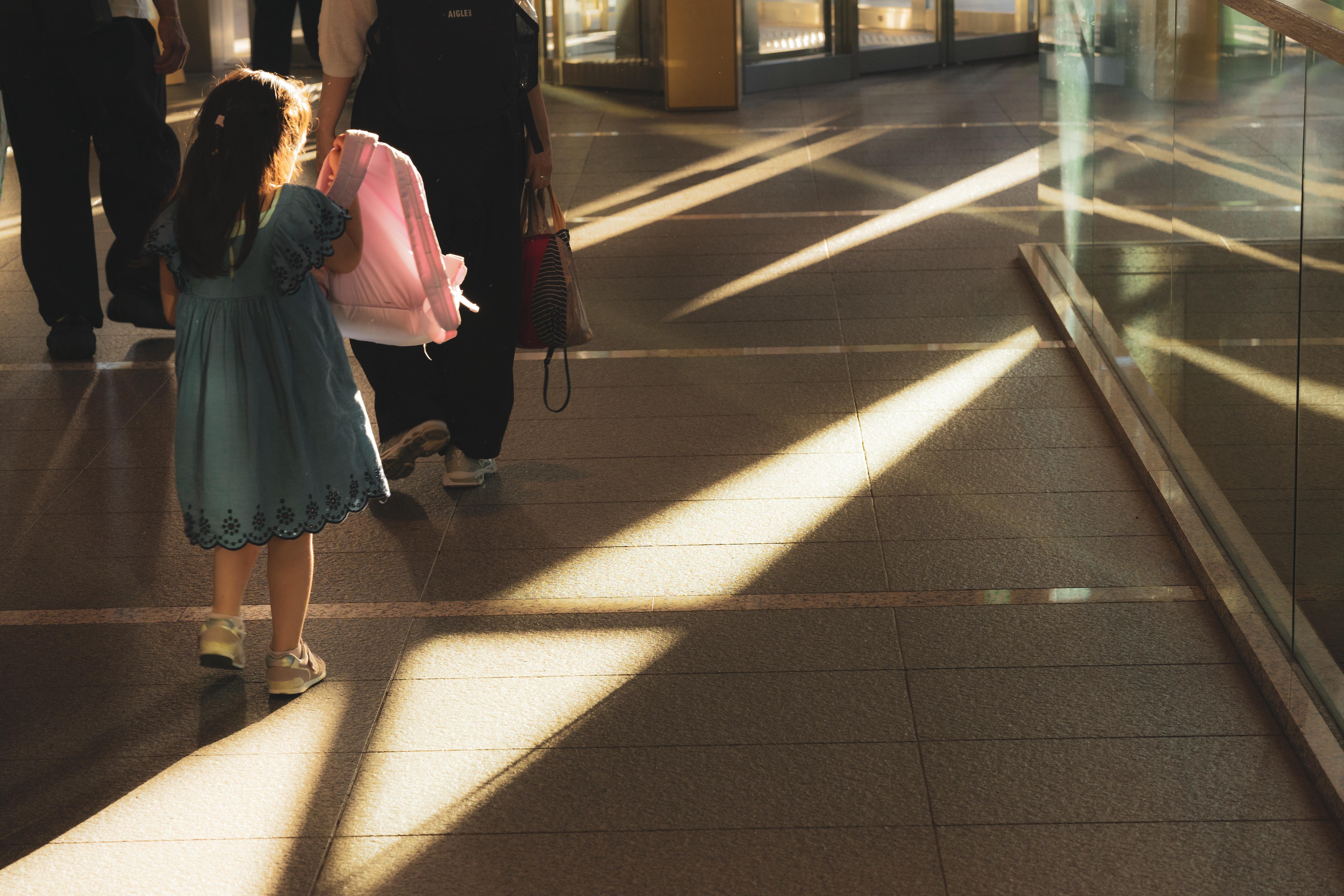 A young girl in a blue dress walks with an adult holding a pink item, possibly a bag, in a sunlit corridor. Strong diagonal sunlight creates patterns on the tiled floor. Glass walls on the right reflect the light, and the corridor appears to lead towards large windows.