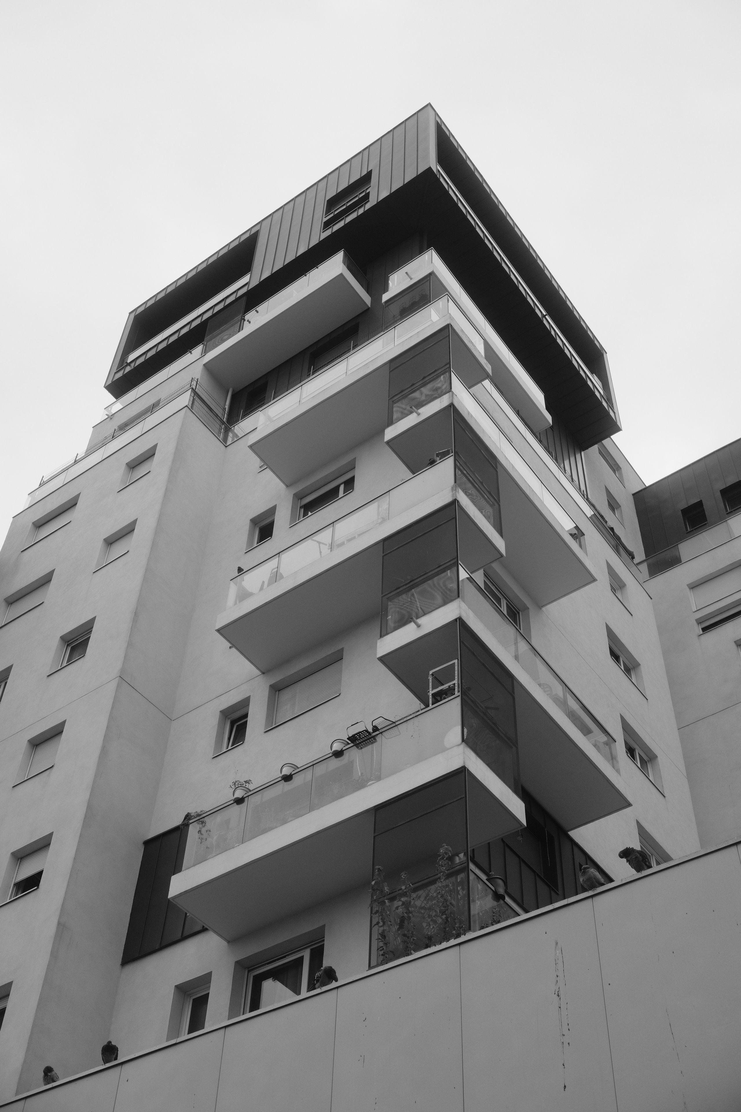Photo verticale en noir et blanc. Contre plongée d’un immeuble moderne blanc, dont le dernier étage est noir