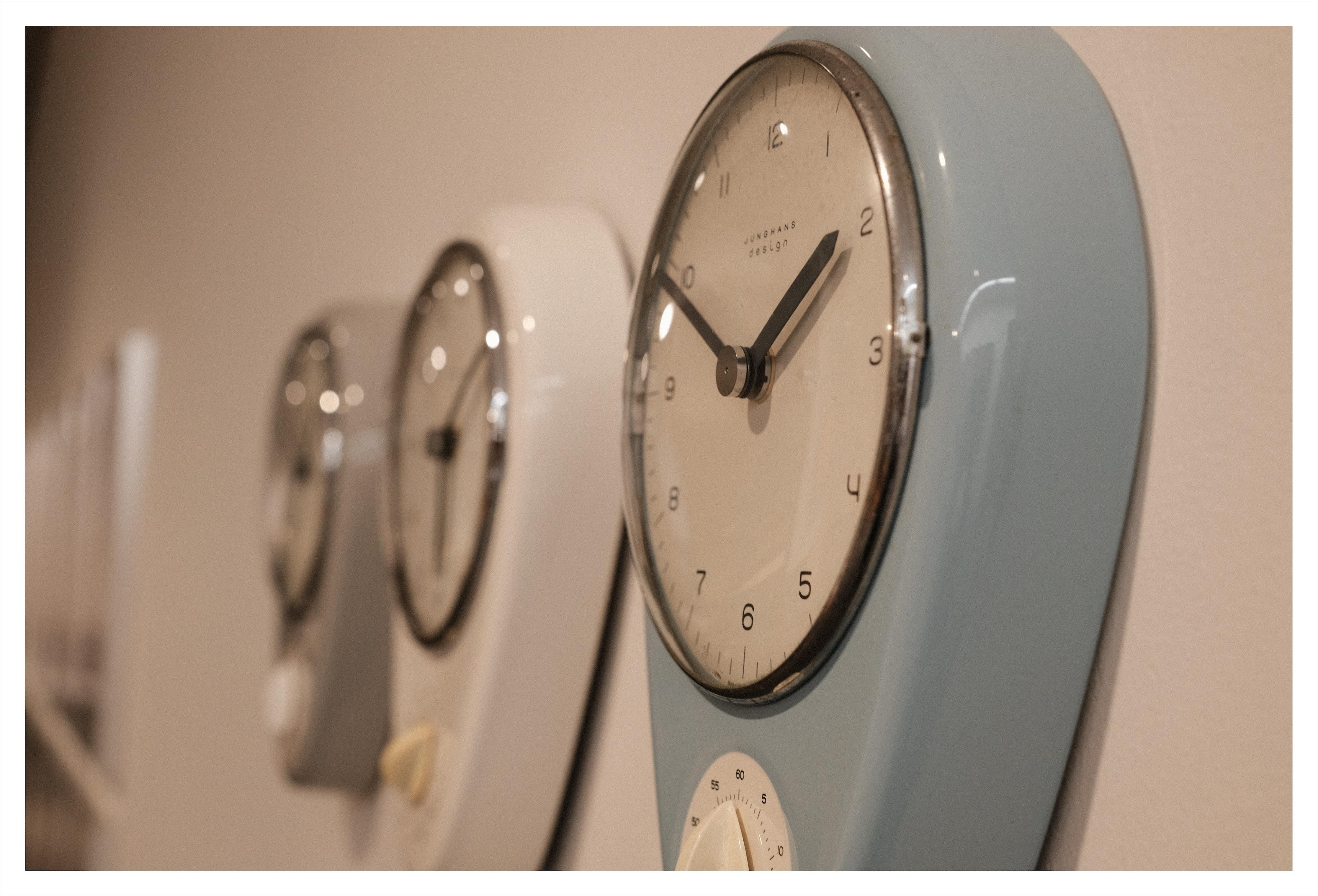 Side angle view of three wall-attached vintage kitchen clocks. In the foreground a baby-blue model, in the midground in white and in the out-of-focus background a light gray model.