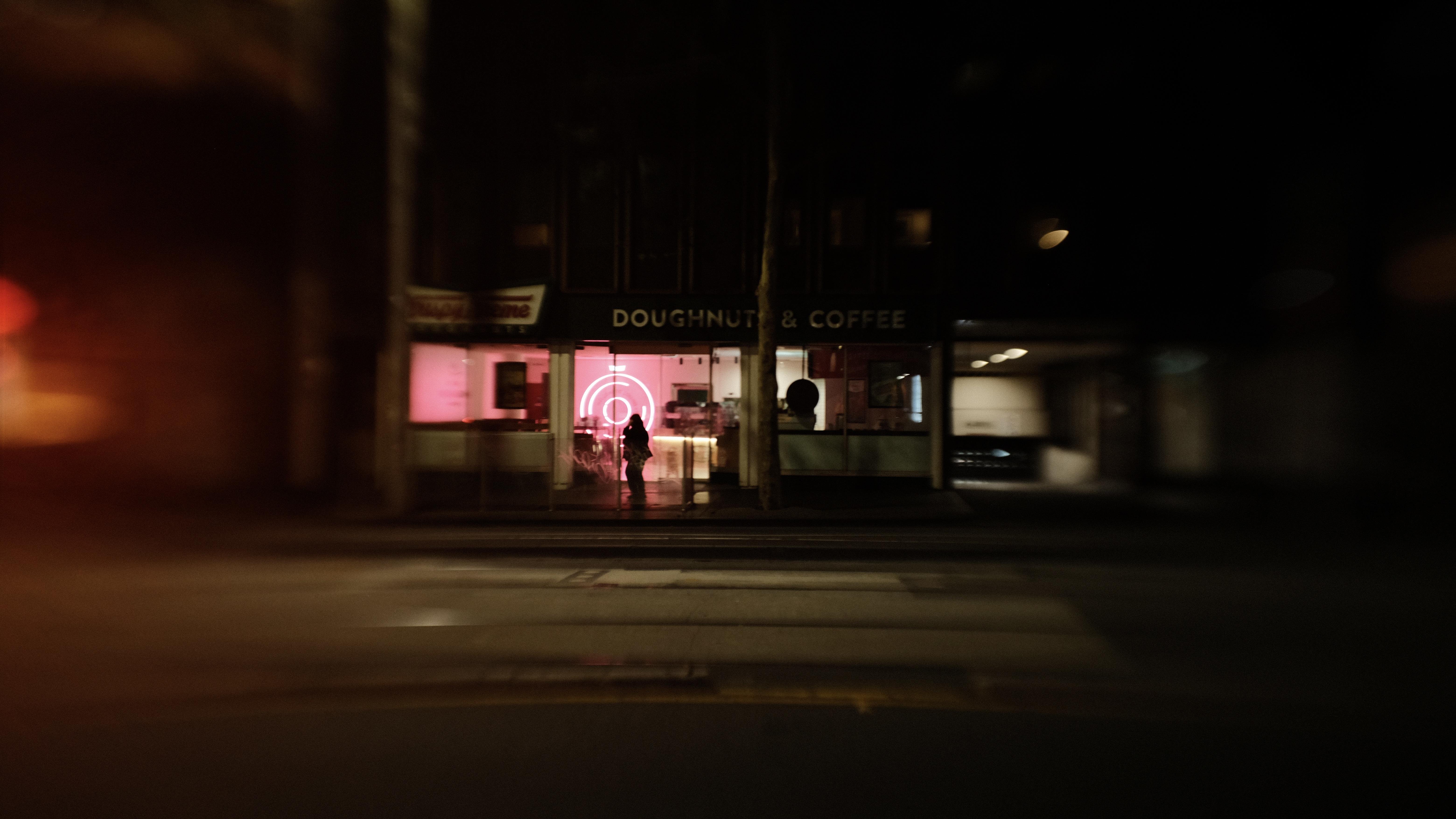 Nighttime street view of a storefront labeled “DOUGHNUTS & COFFEE,” with a pink-lit window and circular neon sign inside; a silhouetted person stands near the entrance behind the glass, and the road with lane markings runs across the foreground.