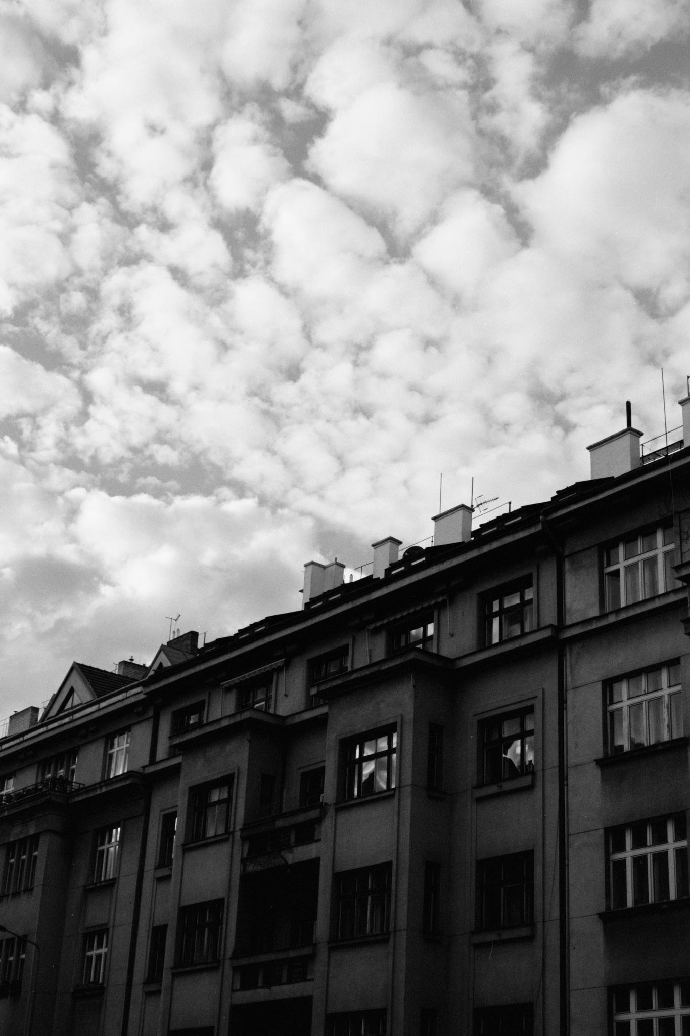 Black-and-white low-angle view of an apartment building facade with rows of windows and rooftop chimneys, beneath a sky filled with dense, puffy clouds.