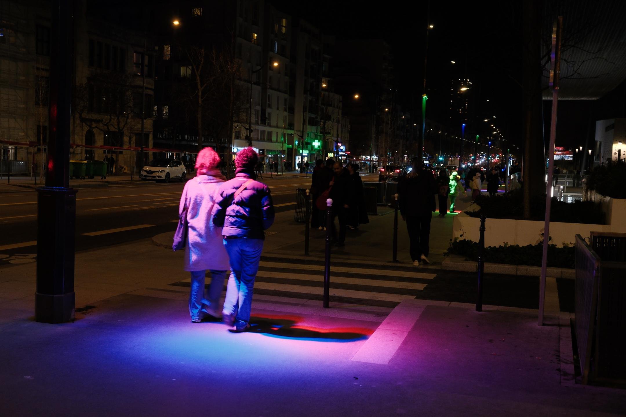 Photo horizontale de nuit. 2 personnes de dos marchant dans la rue et passant sous une lumière bi-colore bleu et rose formant un cercle sur le trottoir