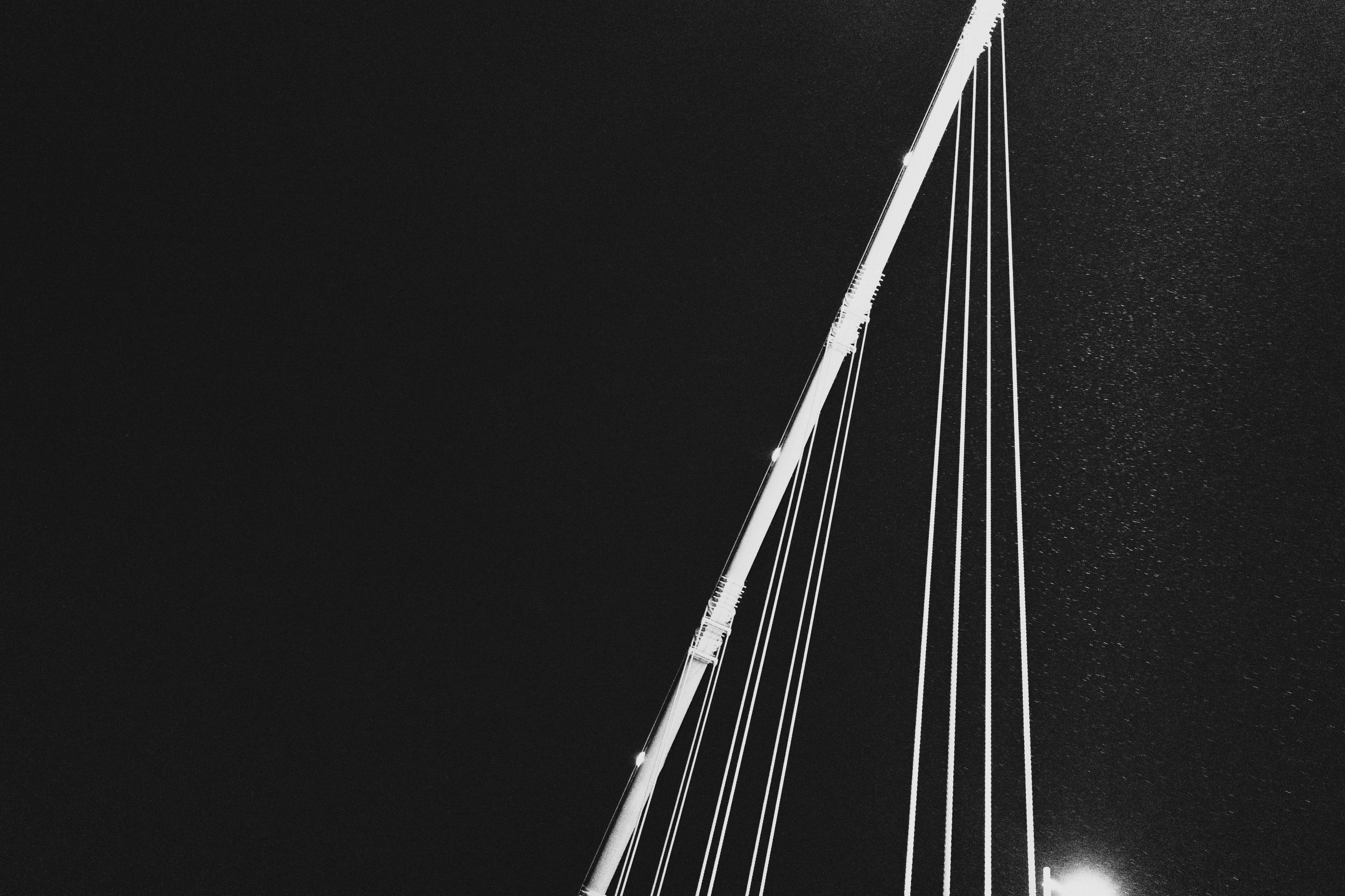 Nighttime view of a suspension bridge's cables and support mast, illuminated against a dark sky. The cables are diagonally aligned from the bottom right to the top left, creating geometric lines. A bright light source is visible at the bottom right corner, casting additional light on the structure. Dust or particles are faintly visible in the air, contributing to the atmospheric effect.