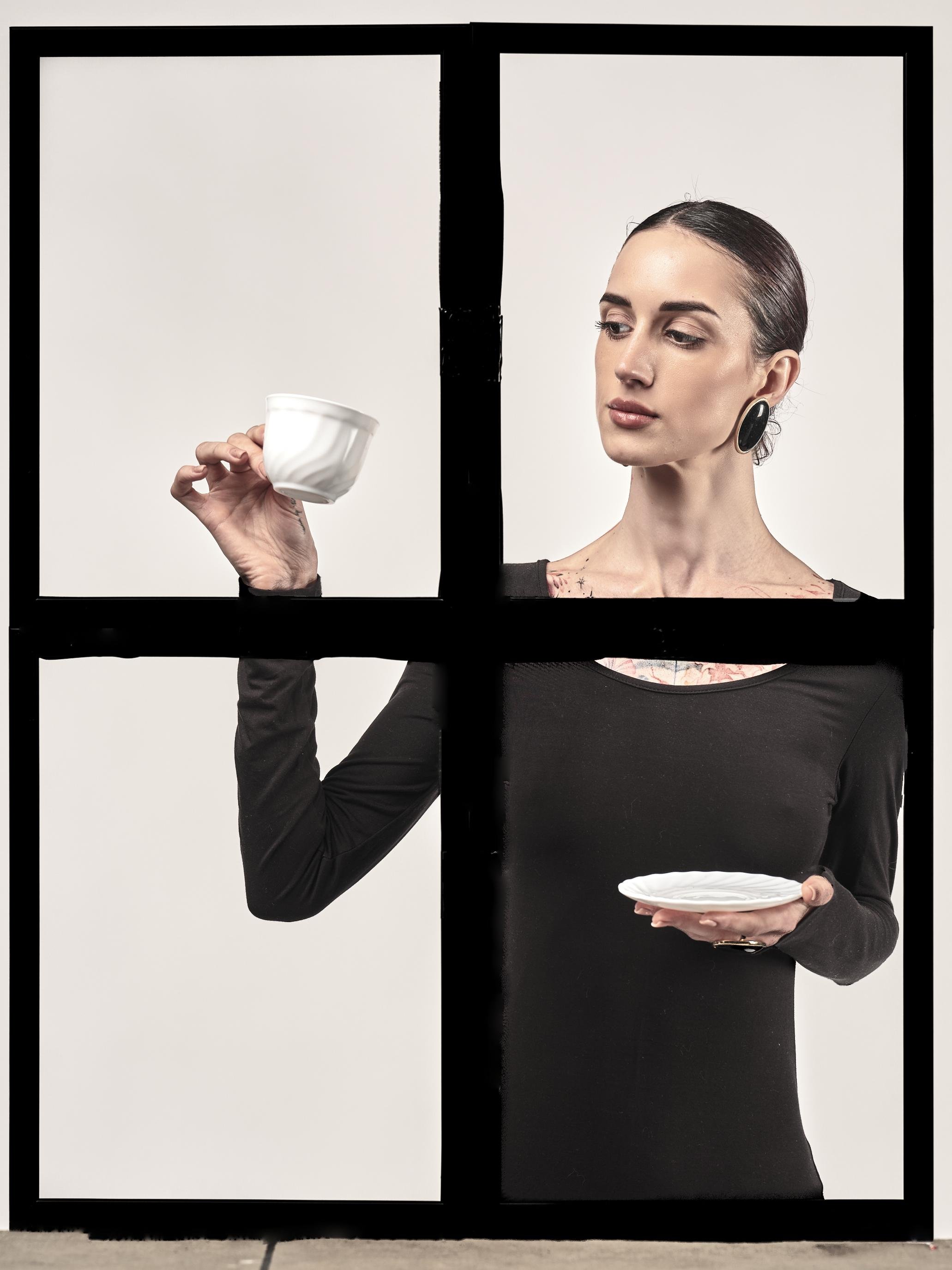 Woman with hair pulled back, wearing a black long-sleeve top and large round earrings, holds a white teacup in her raised left hand and a white saucer in her right, framed by thick black crossbars resembling a window against a plain light background.