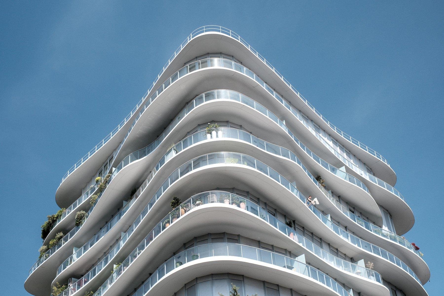Photo horizontale. Contre plongée sur le haut d’un immeuble à l’architecture moderne. Blanc, avec une rambarde en verre sur chaque étage pour sécuriser les balcons. Chaque étage est tout en courbe mais pas identique et l’ensemble forme comme un triangle dont on regarde une pointe en face
