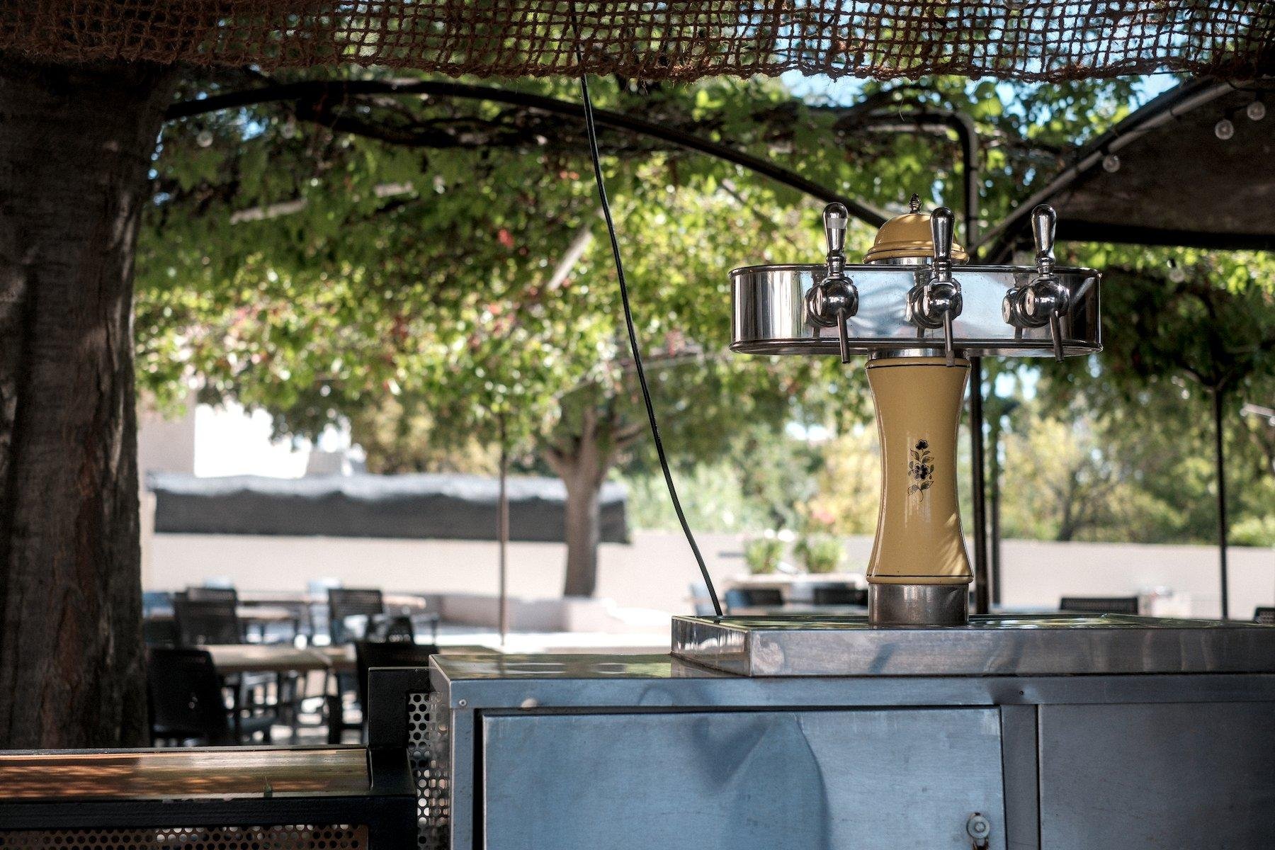Horizontal photo. View on a pressure beer dispenser with 3 hoses. The vertical part is yellow and the horizontal part silver. It stands on a steel furniture, probably a cupboard with the glasses. Behind, we can see the terrace with empty tables and chairs, below some protection against the sun