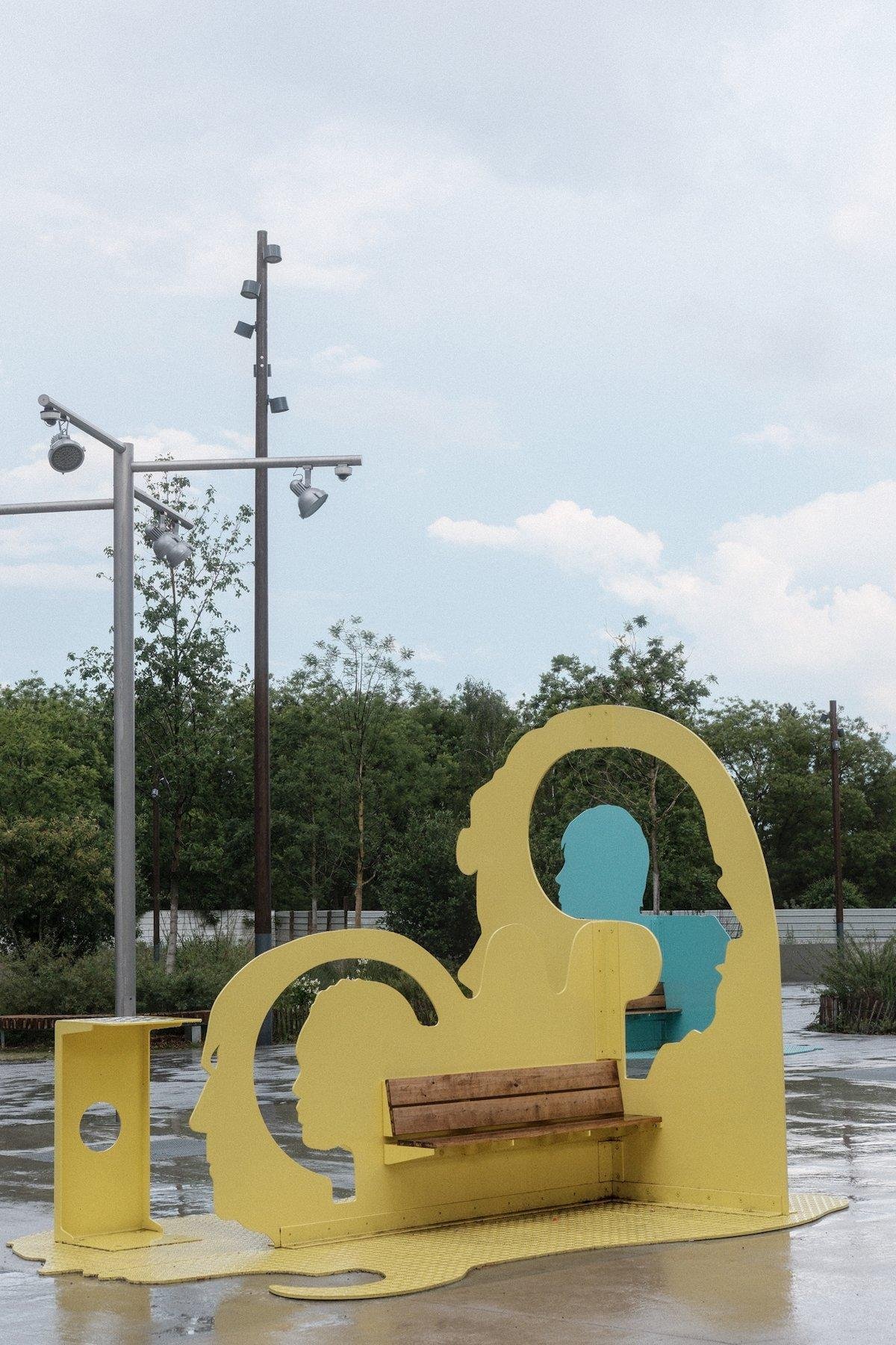 Photo verticale. Au premier plan, un banc dans une structure métallique jaune avec des découpe en forme de silhouettes de visages de profil, et dans l’une d’elle qui regarde à droite, on voit à travers une autre silhouette pleine bleu qui regarde à gauche. En arrière plan, des arbres derrière une haie blanche, le tout sous un ciel bleu post pluie, comme en témoigne le sol mouillé