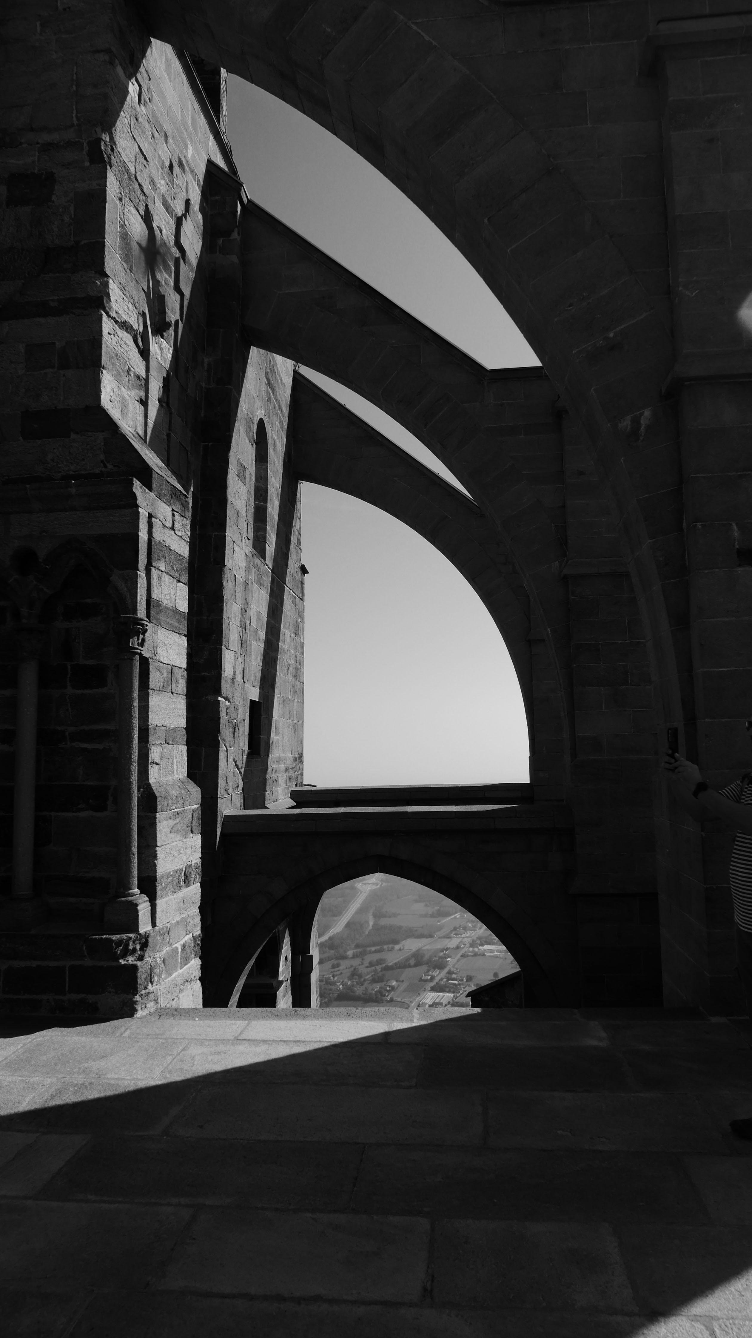 This image depicts an architectural scene featuring stone arches, likely from a historical or Gothic-style structure. The stonework and the shadow play suggest it might be a part of a cathedral or an old building. There is also a person on the right taking a photograph. The view through the arches reveals a distant landscape.