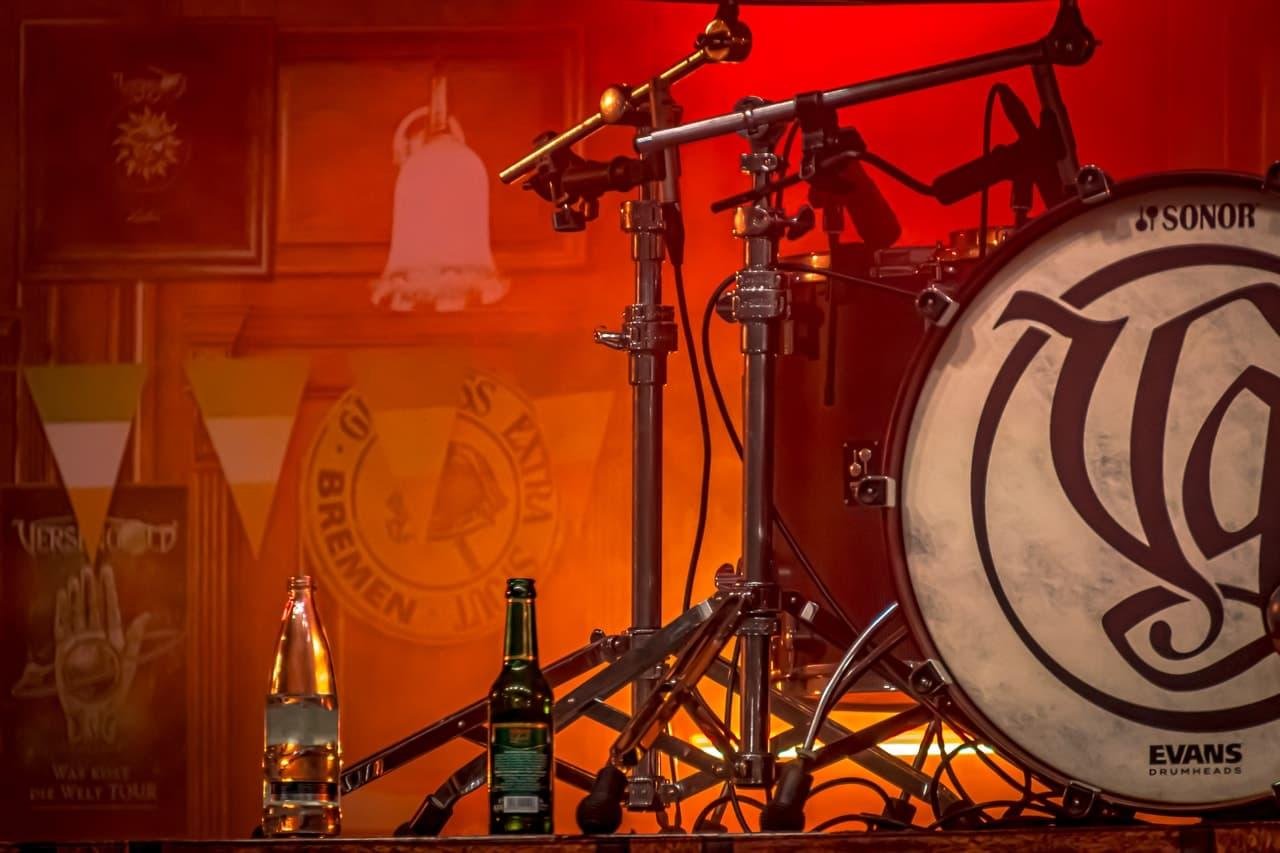 Drum set with a Sonor bass drum and Evans drumhead logo, positioned on a stage with a red-orange backlight. Two bottles, one clear and one green, sit on the floor in front of the drum. Background features a wooden wall with framed art, banners, and various decorations.