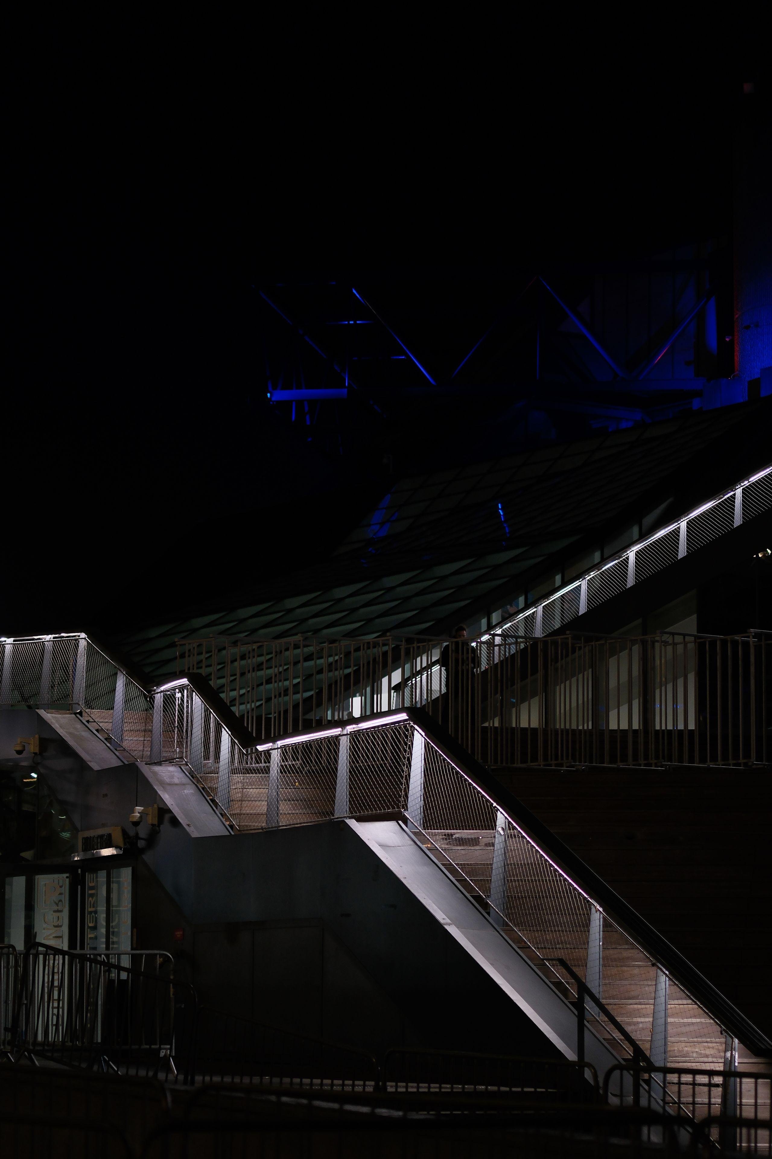 Photo format portrait, de nuit, montrant un escalier illuminé par des LED blanches sous la rampe d’escalier, commençant en bas à droite, pour monter jusqu’au milieu gauche et repartir vers la droite. Presque tout est noir sinon, et un halo de lumière bleu sur le tiers haut de la photo