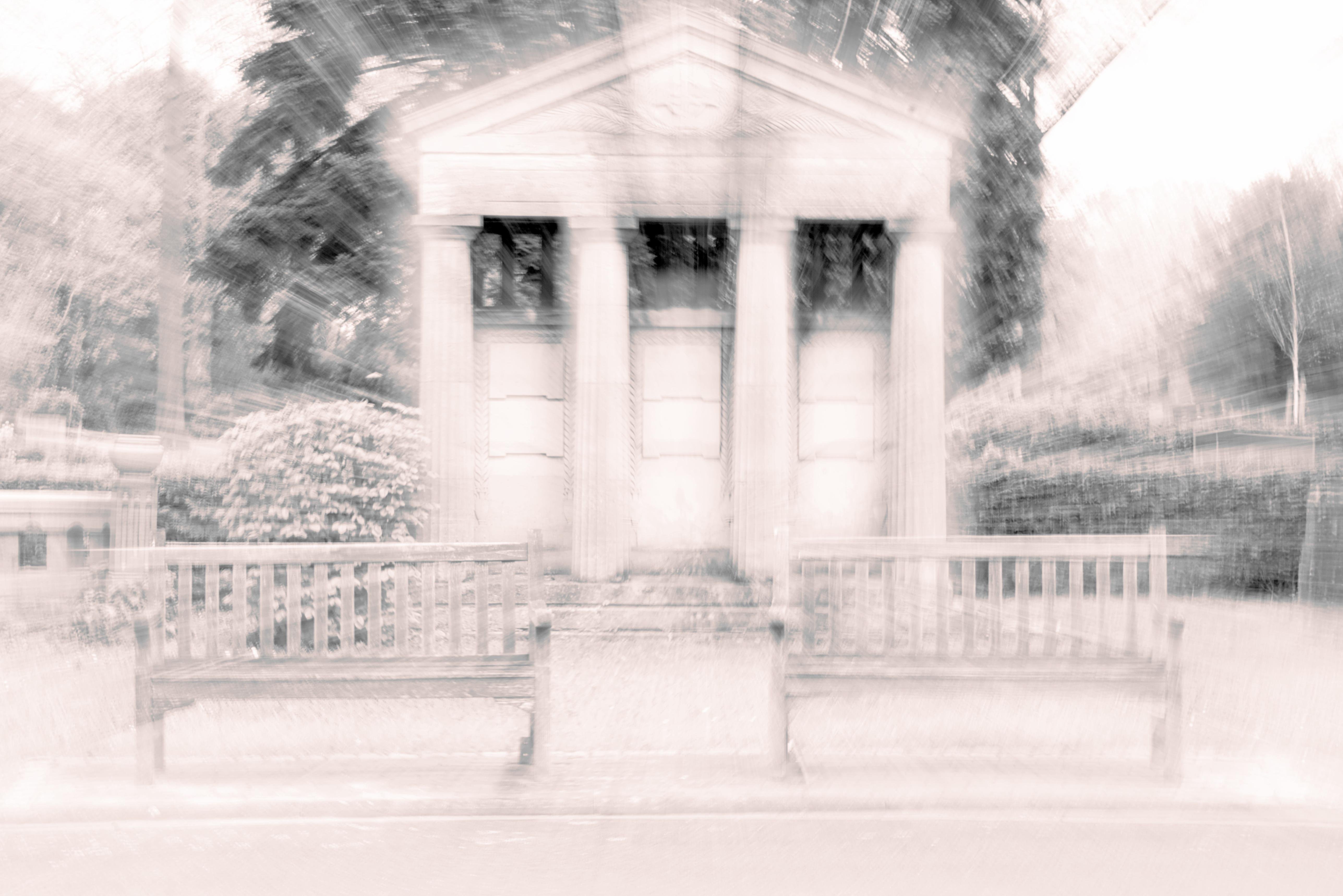 The image appears to be an artistic, blurred photograph of a classical building structure, resembling a mausoleum or a memorial with columns. There are two benches in the foreground, and the background includes trees and possibly a wall or hedge. The blurring effect gives it an abstract, surreal appearance.