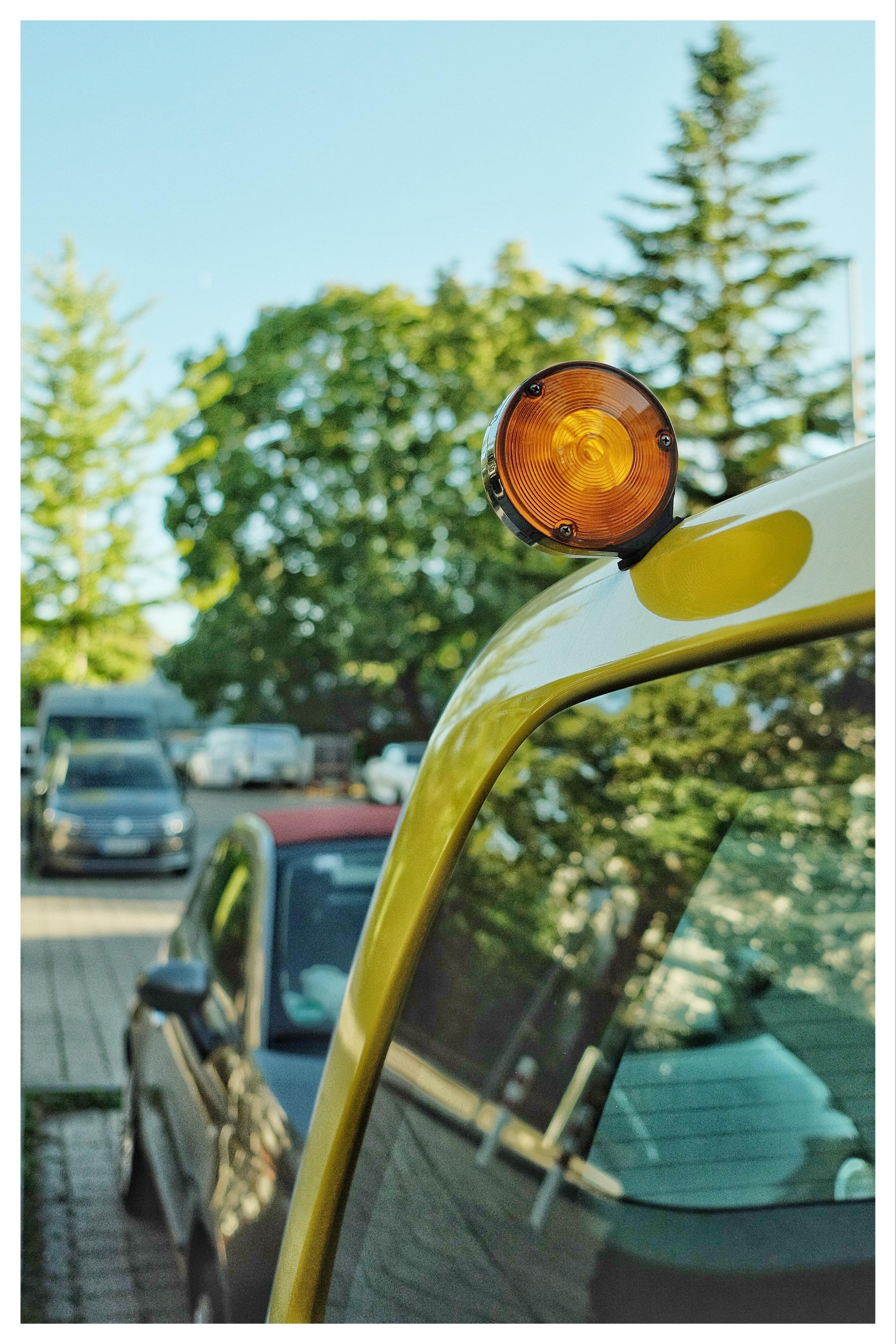 Close-up of an amber round signal light mounted on the roof edge of a yellow vehicle, with parked cars and leafy trees blurred in the background under a blue sky.