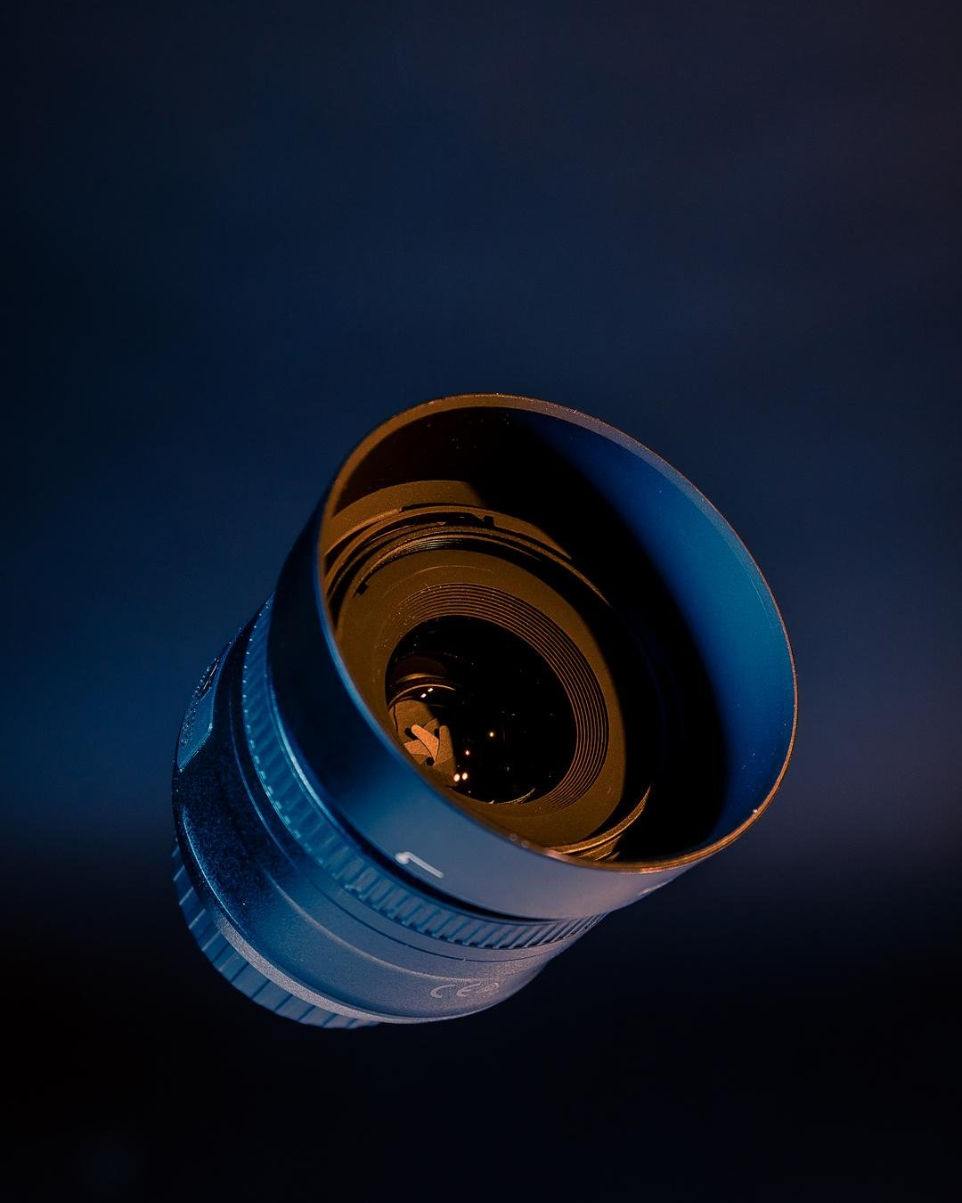 The image shows a camera lens against a dark background. The lens is positioned with the glass facing towards the viewer, allowing you to see the inner elements and the aperture blades. The lighting highlights the details of the lens.