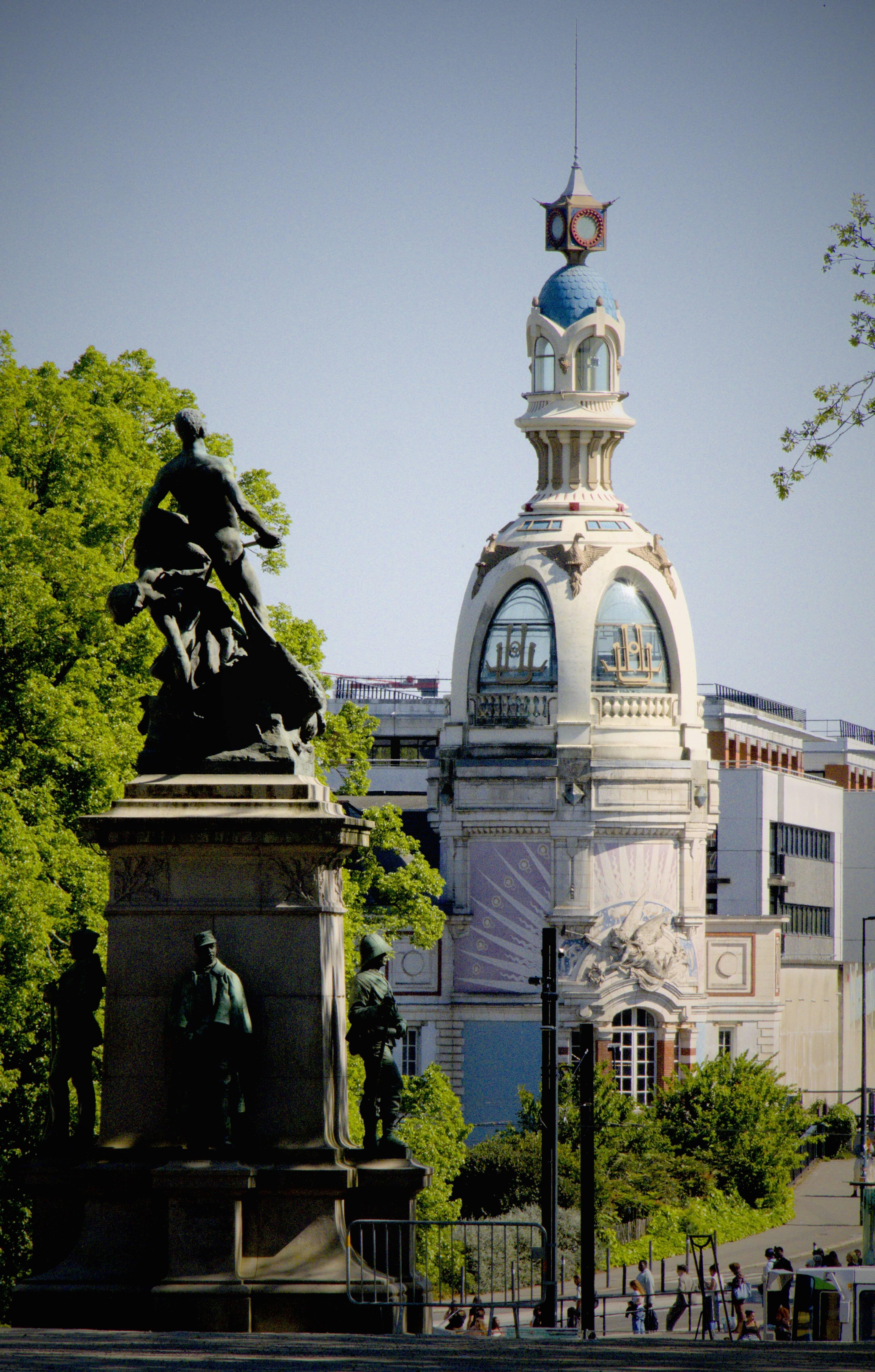 La photographie représente un grand bâtiment orné d'un dôme bleu et de détails architecturaux complexes, avec une horloge au sommet. Le centre culturel Nantais, le Lieu Unique est très reconnaissable. Au premier plan, il y a une statue ou un monument avec plusieurs personnages, plus précisément, le mémorial aux victimes de la guerre de 1870 à Nantes. Il y a des arbres sur la droite et quelques personnes qui marchent en bas à gauche, entre les deux sujets.