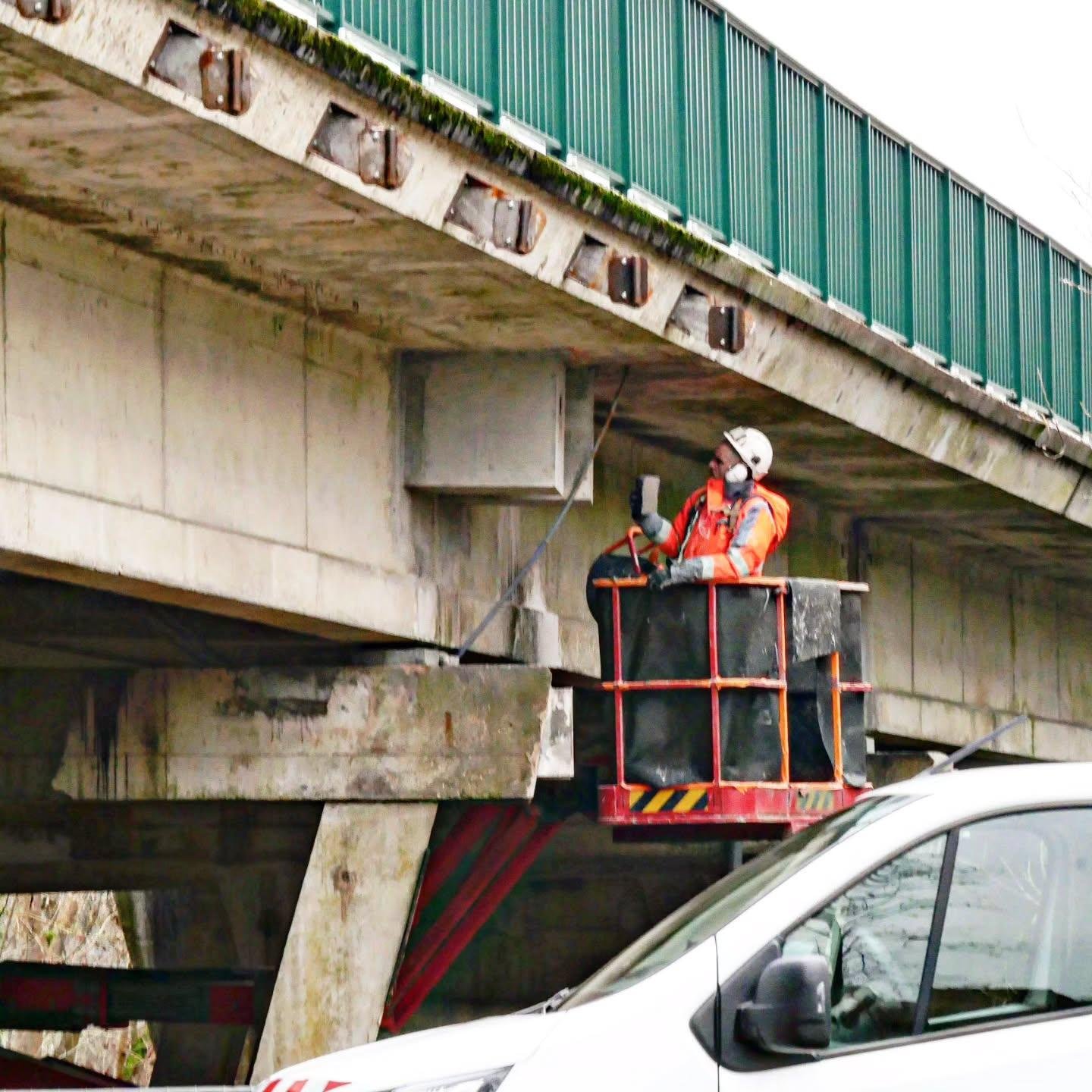 The image shows a worker in a safety lift conducting an inspection or maintenance on the underside of a bridge. The worker is wearing safety gear, including a helmet and an orange reflective jacket. There is a van parked nearby.