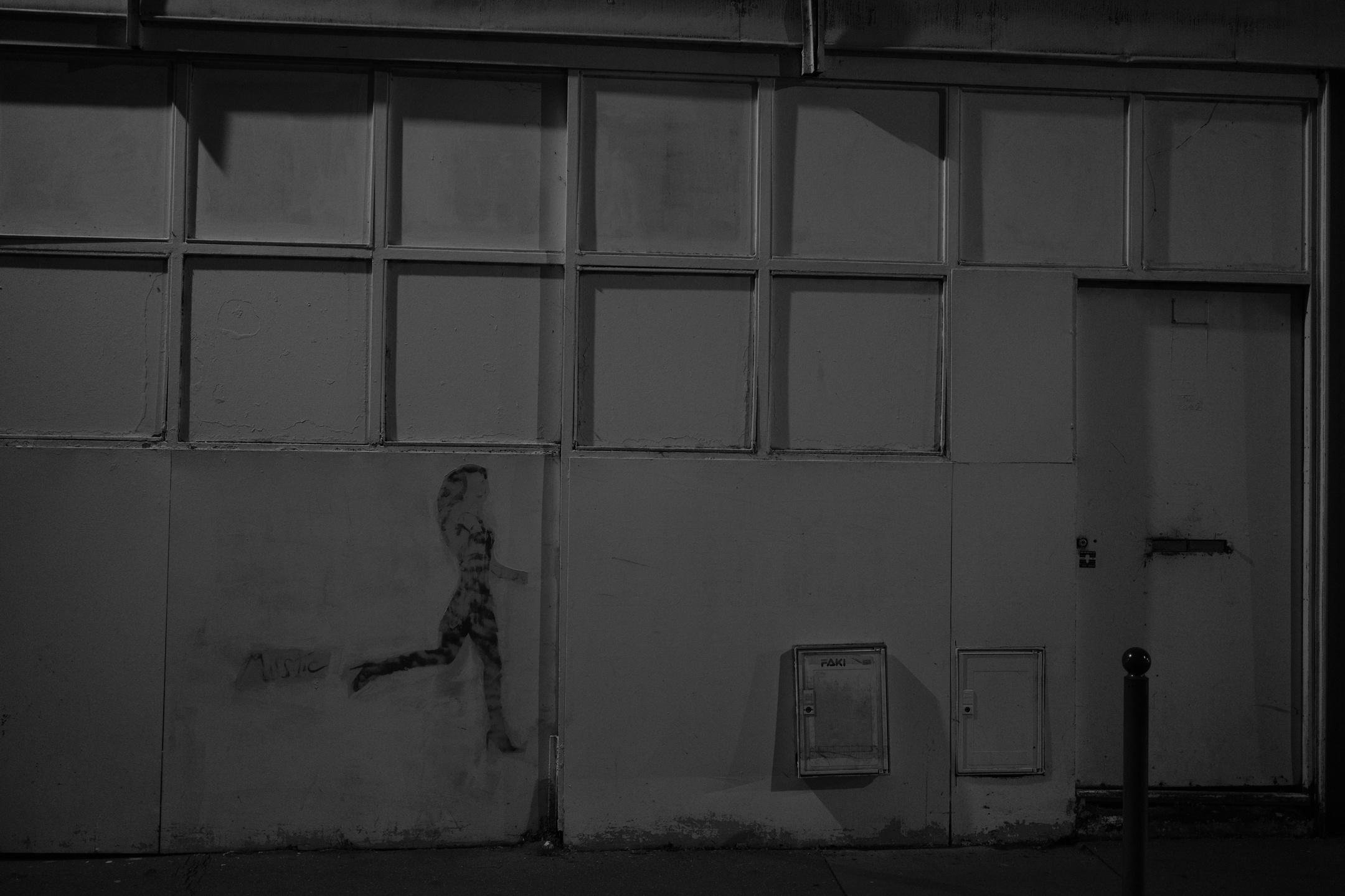Photo format paysage en noir et blanc montrant la façade blanche sale d’un bâtiment qui semble abandonné, les fenêtres étant condamnées. Sur le mur, le graffiti d’une femme qui court, de profile, et qui est bien délavé par le temps