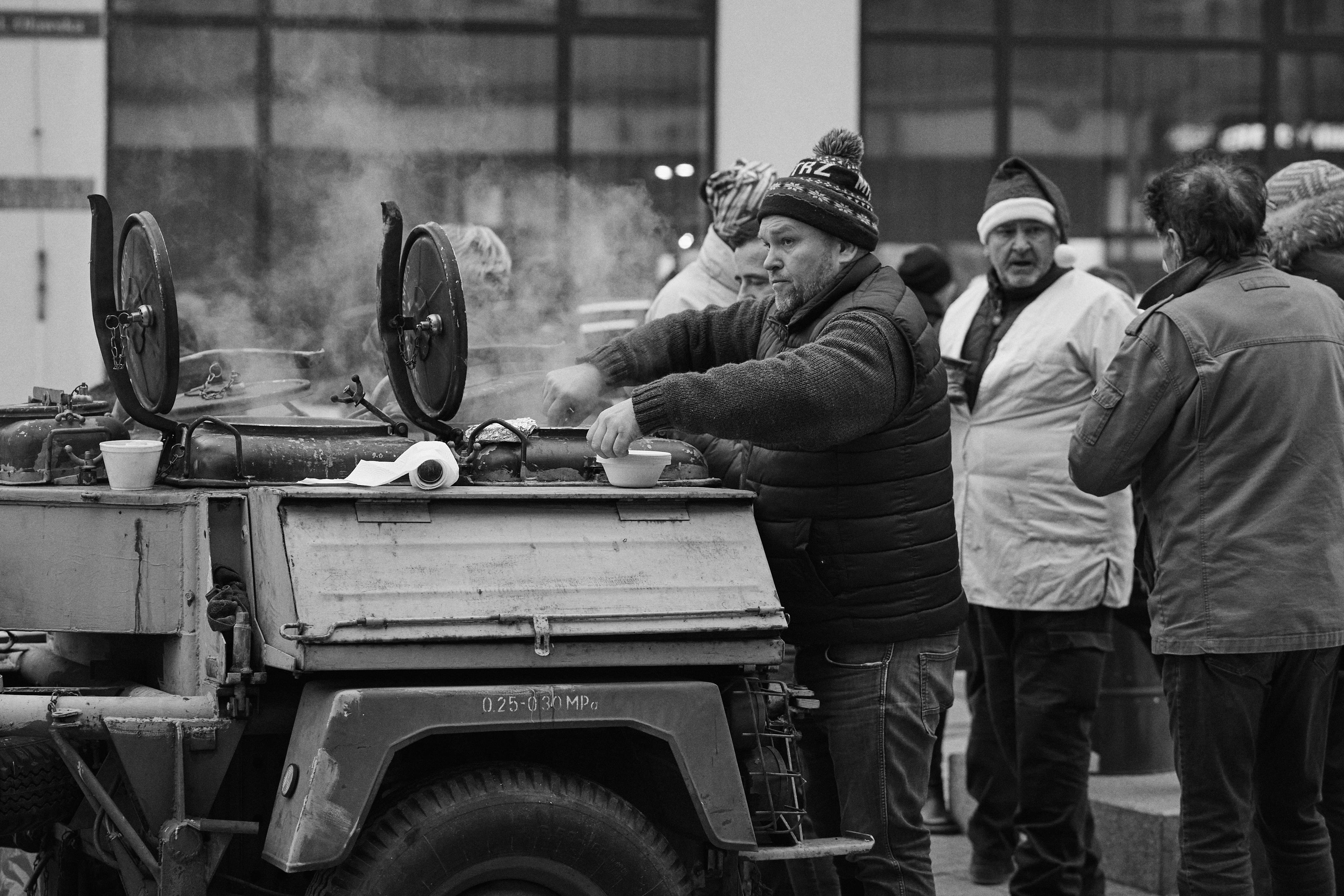 The image shows a group of people gathered around an outdoor food setup. A man is serving food from a mobile cooking unit, which appears to be an old military-style field kitchen. Steam is rising from the cooking equipment, indicating that it's being used to prepare or serve hot food or drinks. The people are dressed warmly, suggesting it's a cold day.