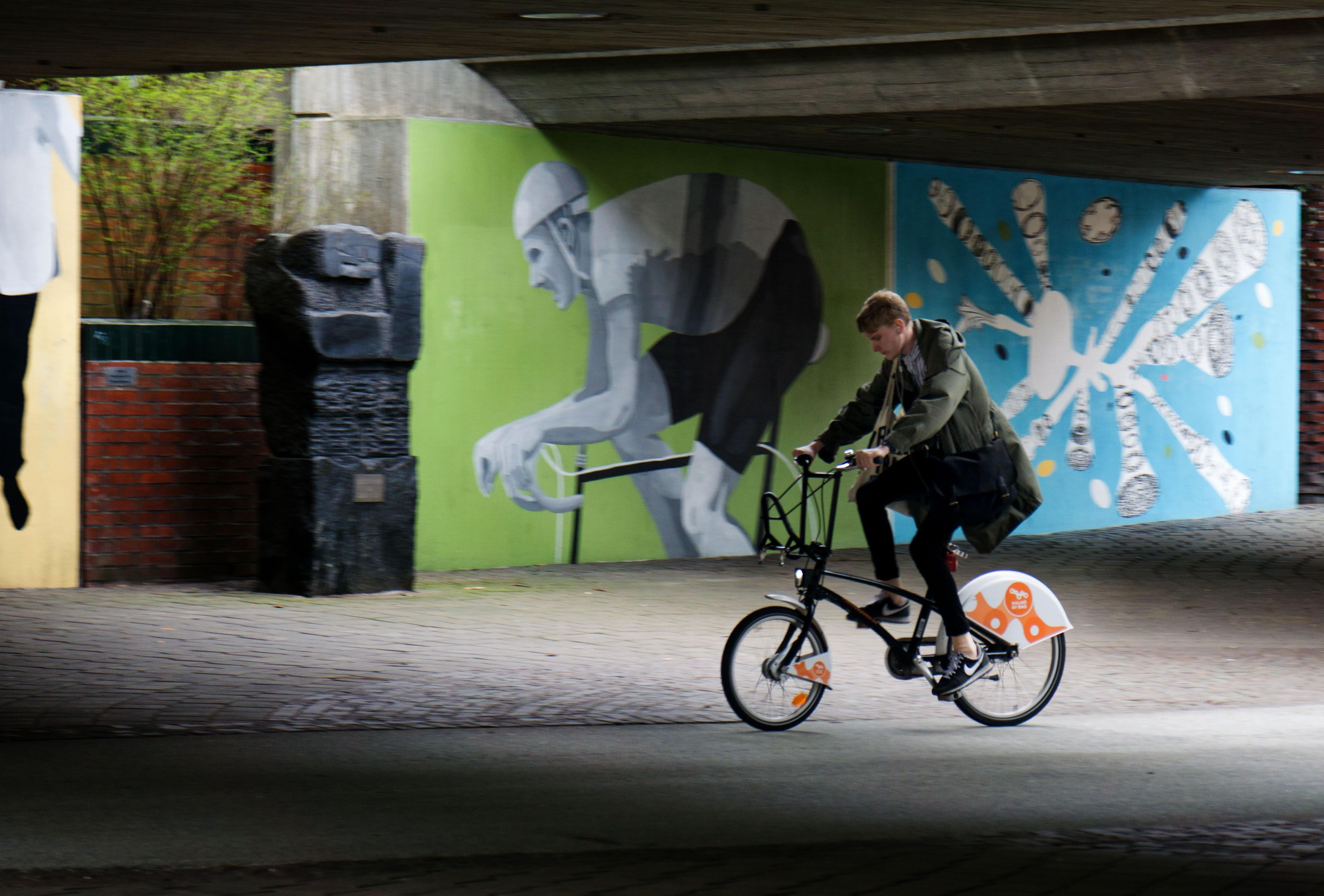 Person riding a bicycle under a concrete overpass, passing large wall murals of a crouching cyclist on a green panel and a white abstract shape on a blue panel; a dark stone sculpture stands near a brick wall on the left.