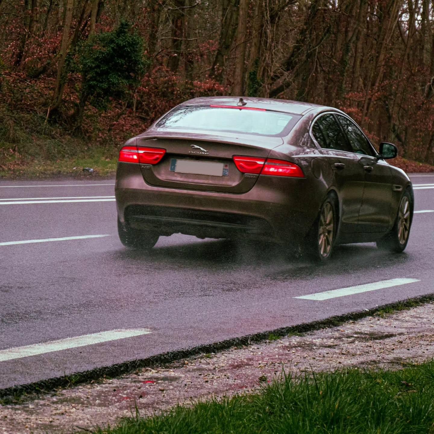 The image shows a car driving on a wet road with trees in the background. The car has visible taillights and exhaust visible, indicating movement.