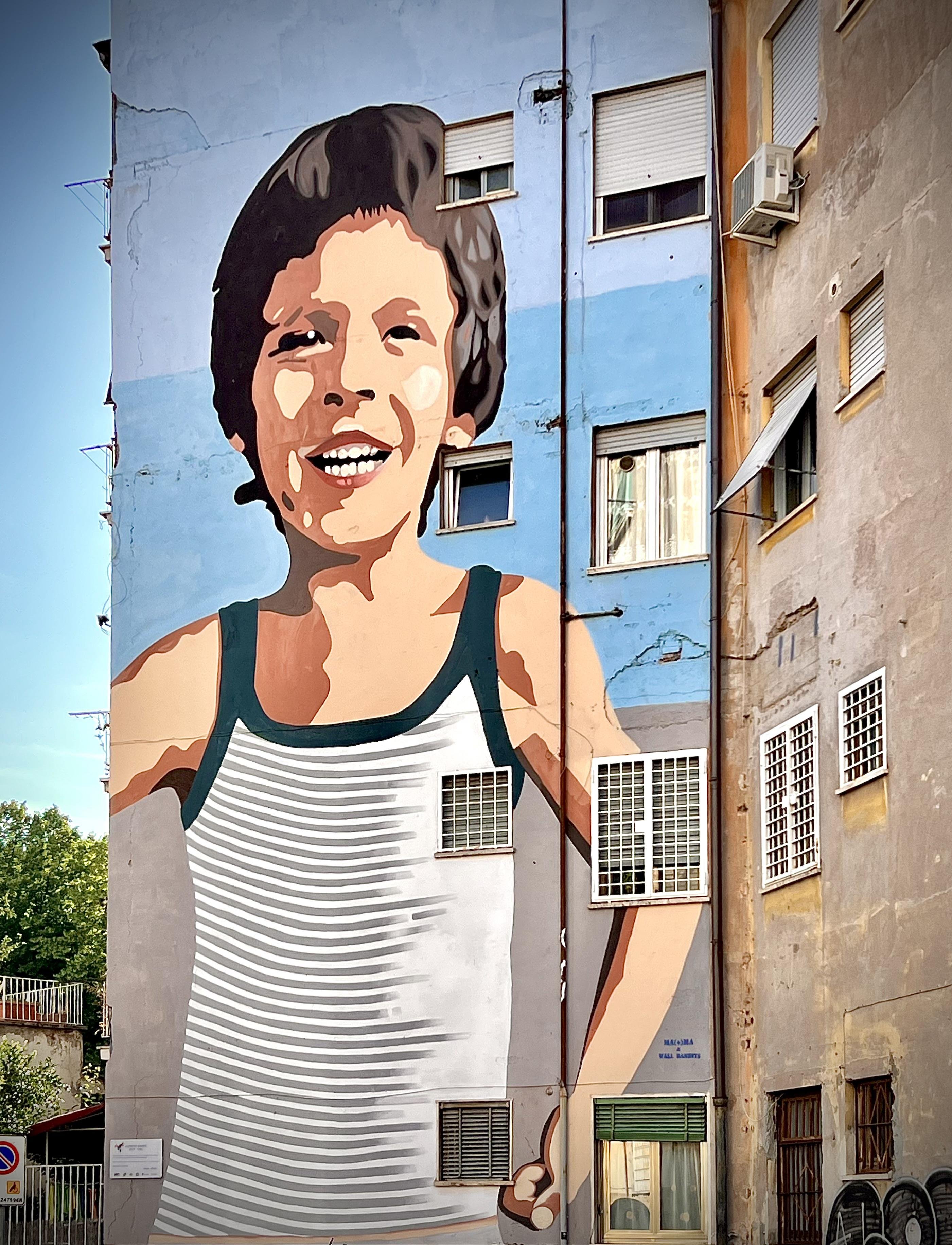 Murale dipinto sul lato di un edificio. Presenta una raffigurazione stilizzata di un bambino sorridente che indossa una canottiera a righe. Il murale è grande e copre più piani dell'edificio, integrandosi con le finestre e gli elementi architettonici.