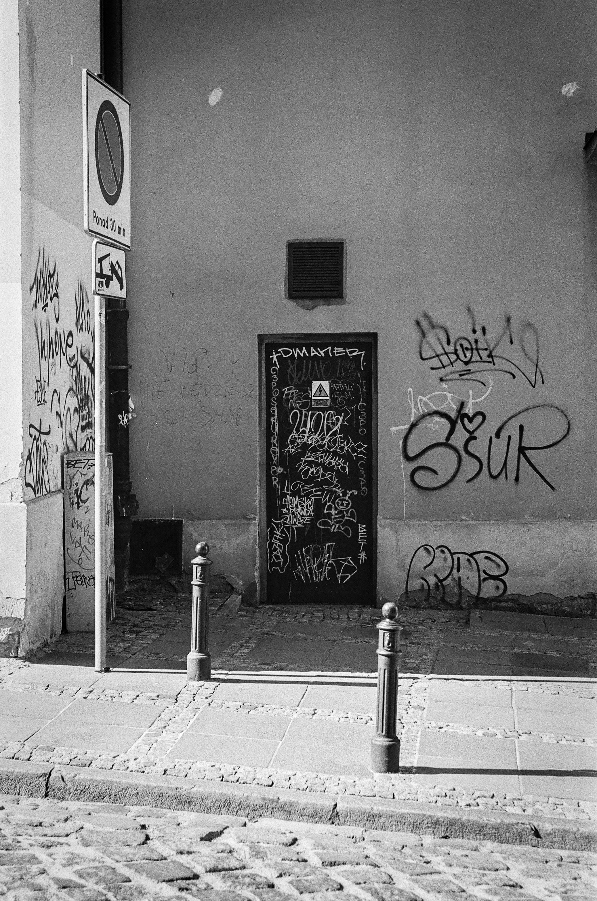 B&W film photo of a street corner in the shade, with an entry to an electrical transformer and walls covered in graffiti