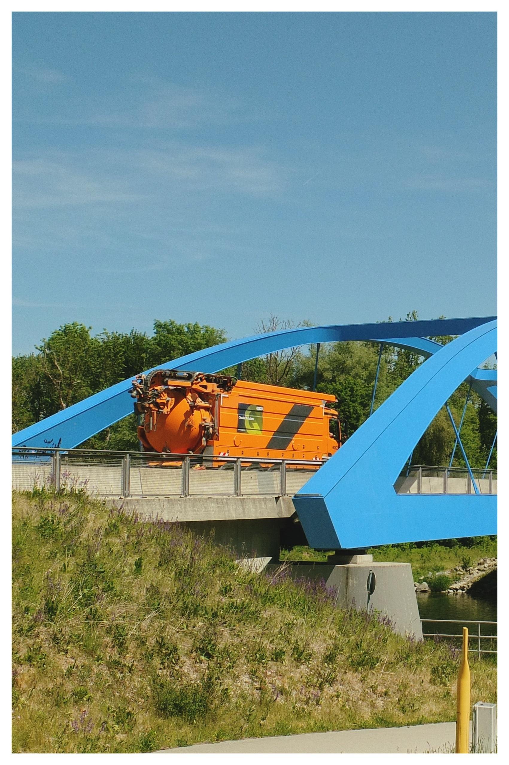 Orange industrial railcar on a blue arched bridge over a river, with green trees and grassy embankment in the foreground under a clear sky.