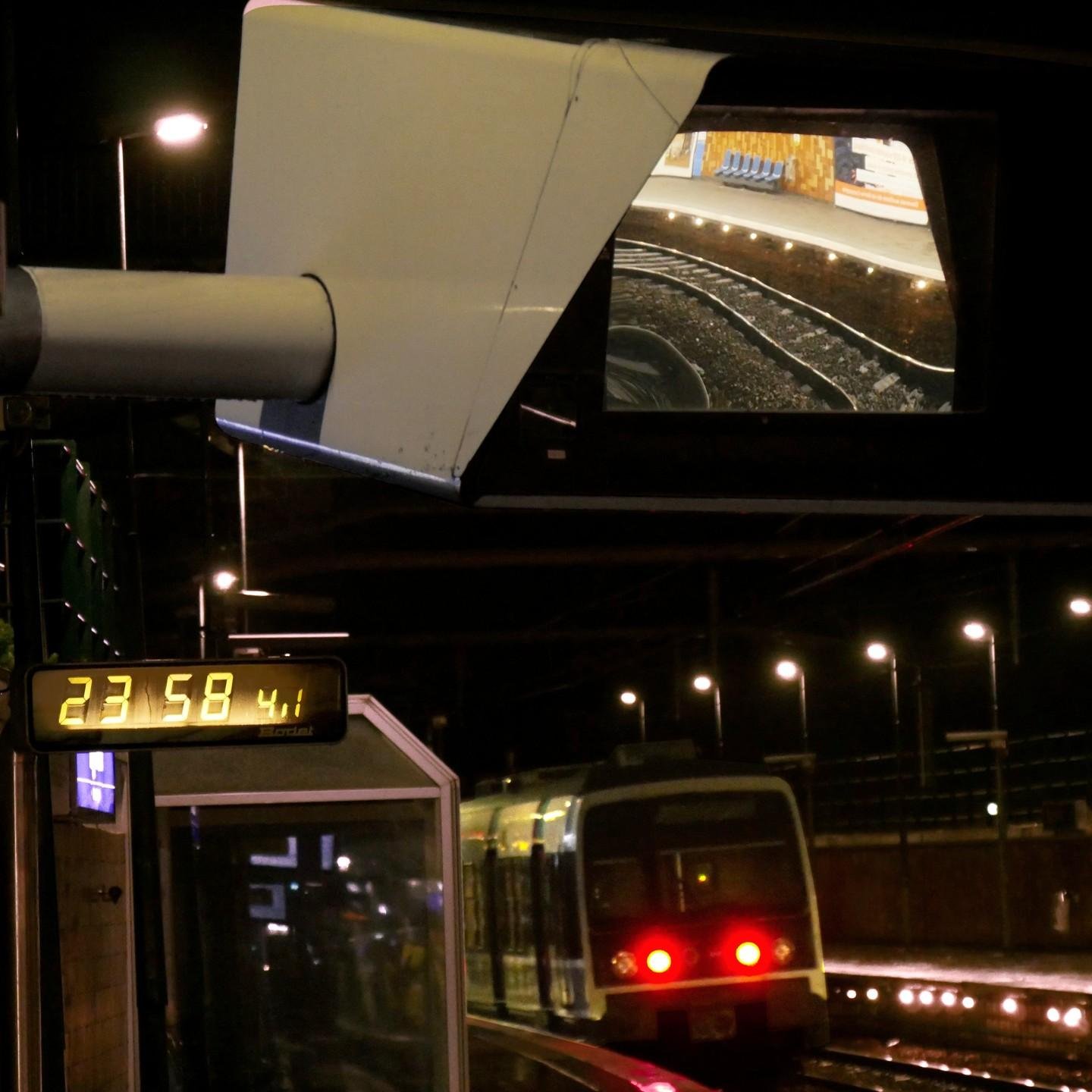 The image shows a train platform at night. A train is running away, and there are various lights illuminating the area. A prominent feature is a large miror displaying a section of the tracks and platform. Additionally, a digital clock shows the time as 23:58.