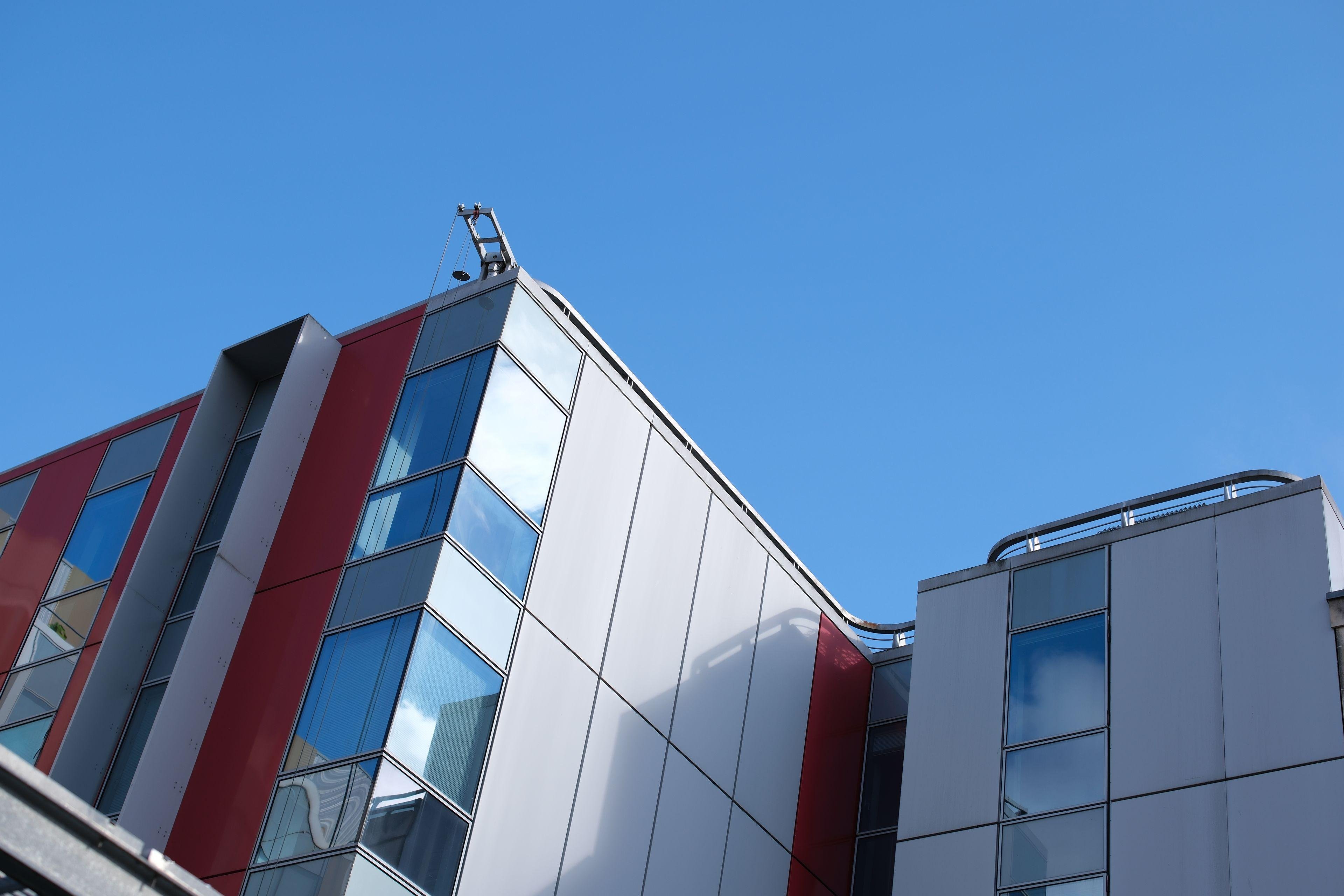 Photo horizontale. Contre plongée sur le haut d’un immeuble moderne blanc aux accents rouge. On voit une rembarde de sécurité le long du bord du toit