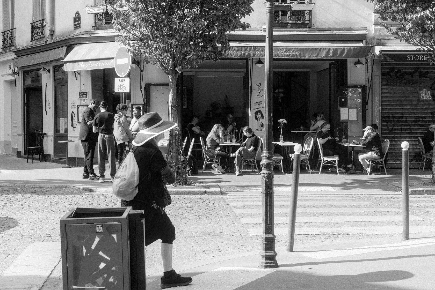 Photo horizontale en noir et blanc. Une dame franchi la marche du trottoir, avec son large chapeau et un sac à dos, en short basket et un haut à manches longues