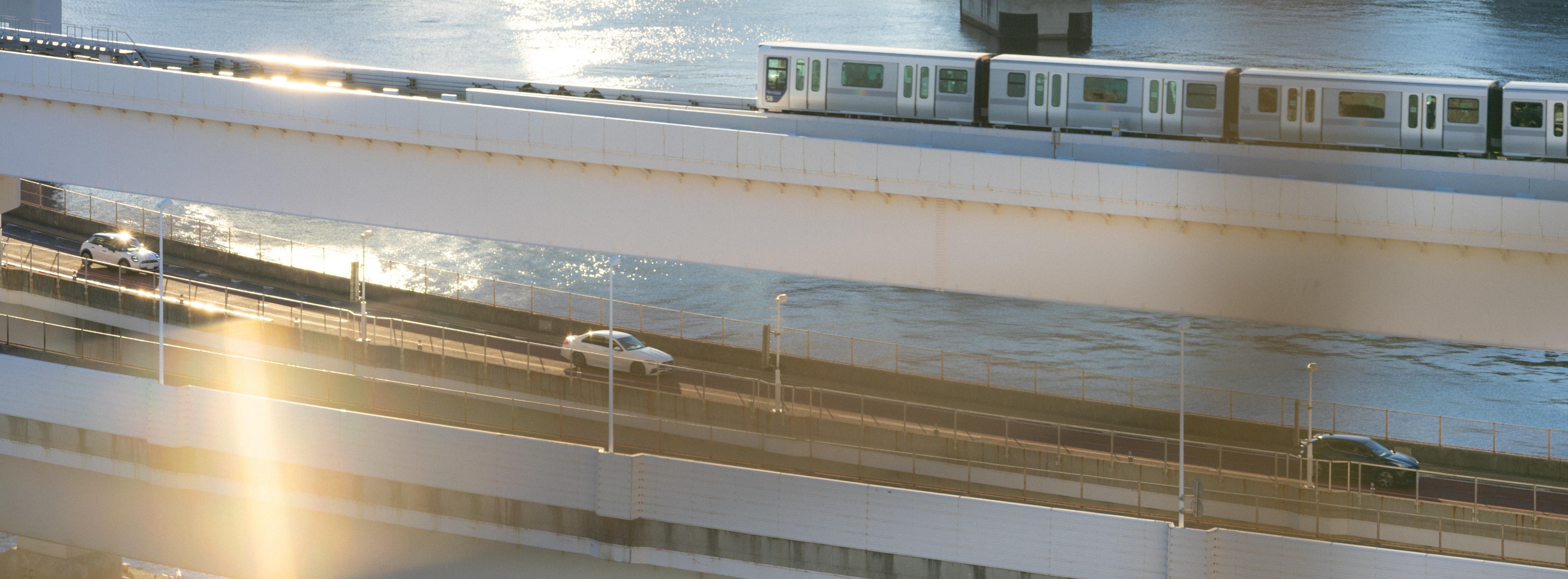 Elevated train traveling on a bridge above a river. Below the train, there is a roadway with two visible cars, separated from the train bridge by another structure. The sun creates a lens flare, casting a warm glow on the scene.