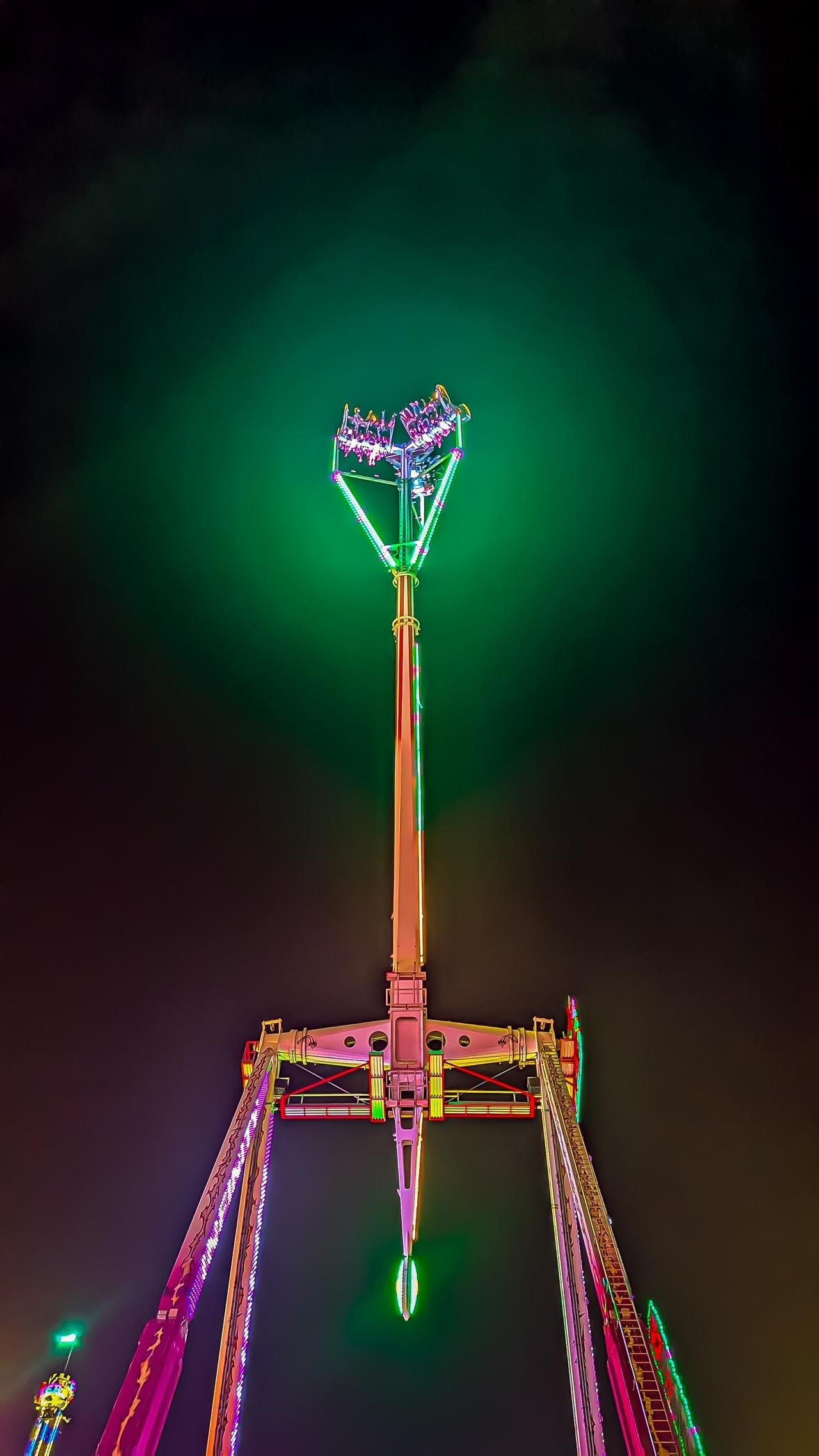 Tall amusement park ride with a central pole and two arms, each holding a gondola with people. Neon lights in green, pink, and yellow illuminate the structure against a dark sky. A bright green glow surrounds the top section.