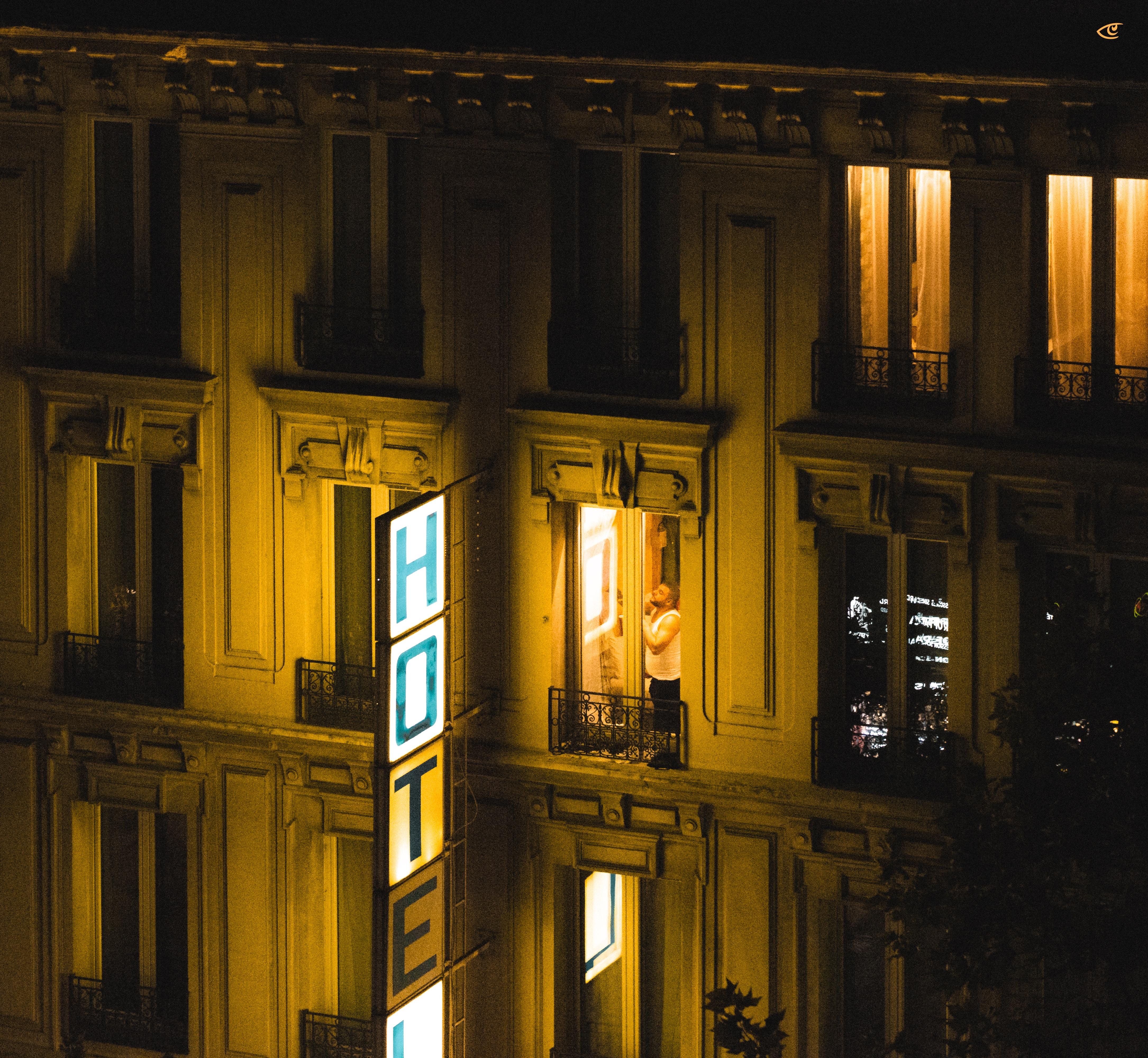 Nighttime view of a multi-story building with a vertical illuminated “HOTEL” sign on the facade; several windows glow with warm light, including one open window where a person stands on a small balcony railing.