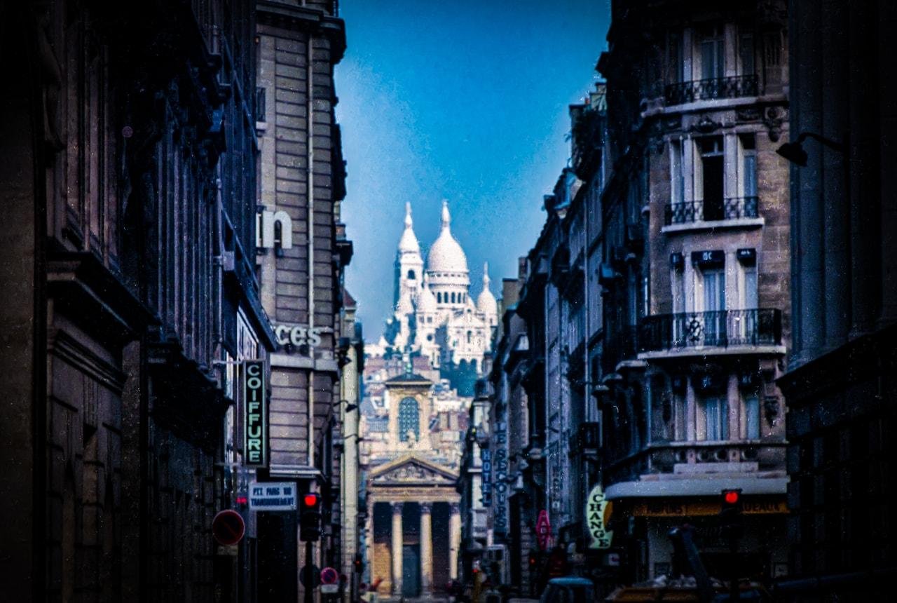 Narrow street in Paris with tall, ornate buildings on either side. In the background, the Basilica of Sacré-Cœur is prominently visible with its white domes against a clear blue sky. Various shop signs in French are visible along the street edges.